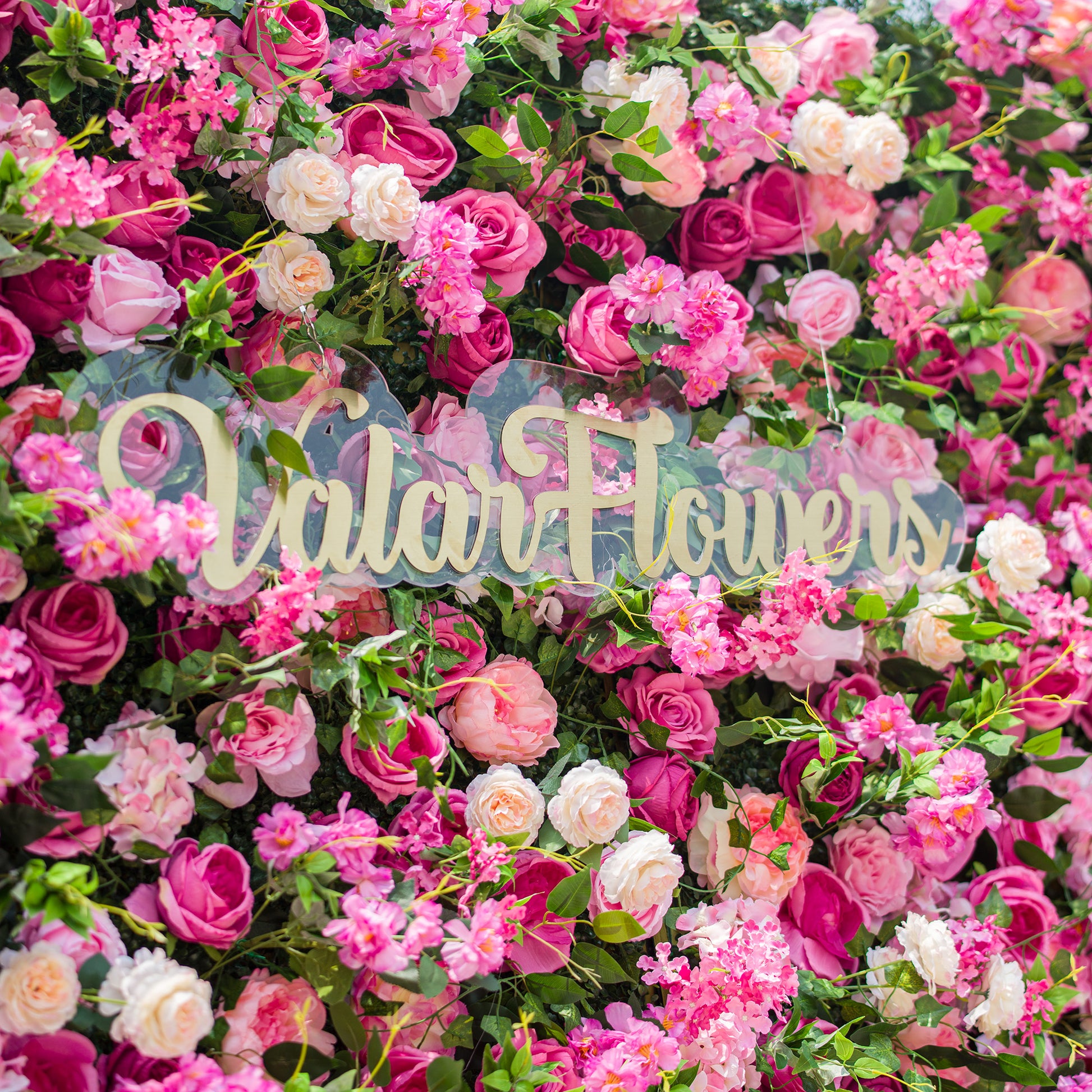 This photo showcases a lush and vibrant floral backdrop densely packed with an array of pink and white roses, along with other assorted pink flowers, all interspersed with rich green foliage. The text "ValarFlowers" in elegant golden lettering stands out prominently. The overall scene exudes a lively, romantic charm, making it ideal for events like garden weddings or spring festivals. 
