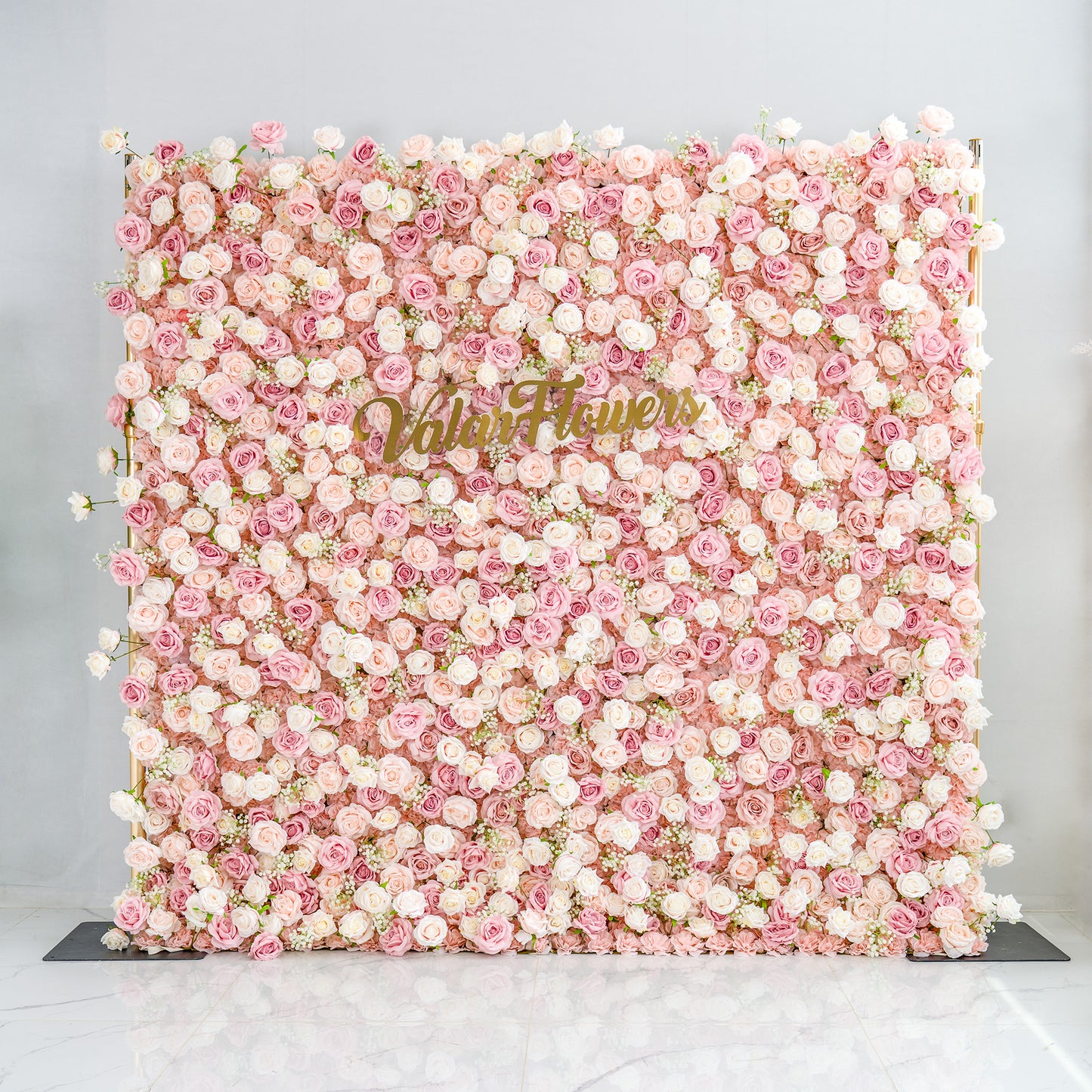 This photo showcases a large floral backdrop densely covered with pink and white roses, interspersed with delicate white baby's breath. The soft pastel color palette creates a romantic and elegant atmosphere. The words "Valar Flowers" in elegant gold lettering stand out prominently. Mounted on a golden frame, it's ideal for weddings or upscale events, exuding a refined, dreamy style. 