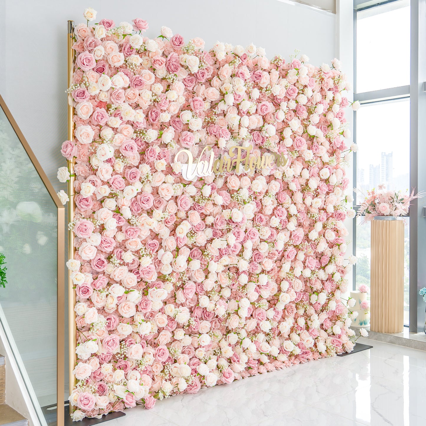 This photo showcases a large floral backdrop densely adorned with pink and white roses, interspersed with delicate white baby's breath. The soft pastel color palette creates a romantic and elegant atmosphere. The words "Valar Flowers" in elegant gold lettering stand out prominently. Ideal for weddings or upscale events, it exudes a refined, dreamy style. 