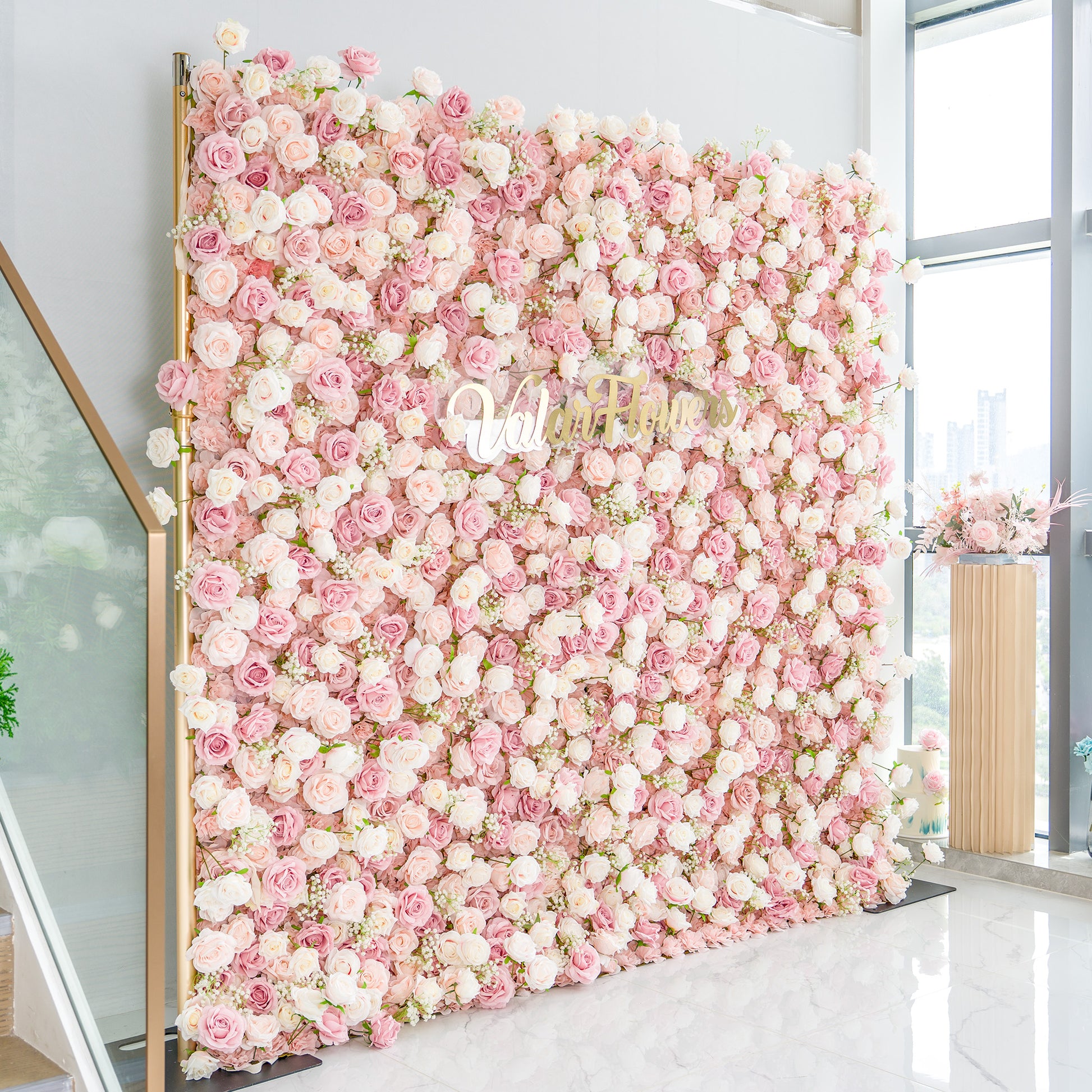 This photo showcases a large floral backdrop densely adorned with pink and white roses, interspersed with delicate white baby's breath. The soft pastel color palette creates a romantic and elegant atmosphere. The words "Valar Flowers" in elegant gold lettering stand out prominently. Ideal for weddings or upscale events, it exudes a refined, dreamy style. 