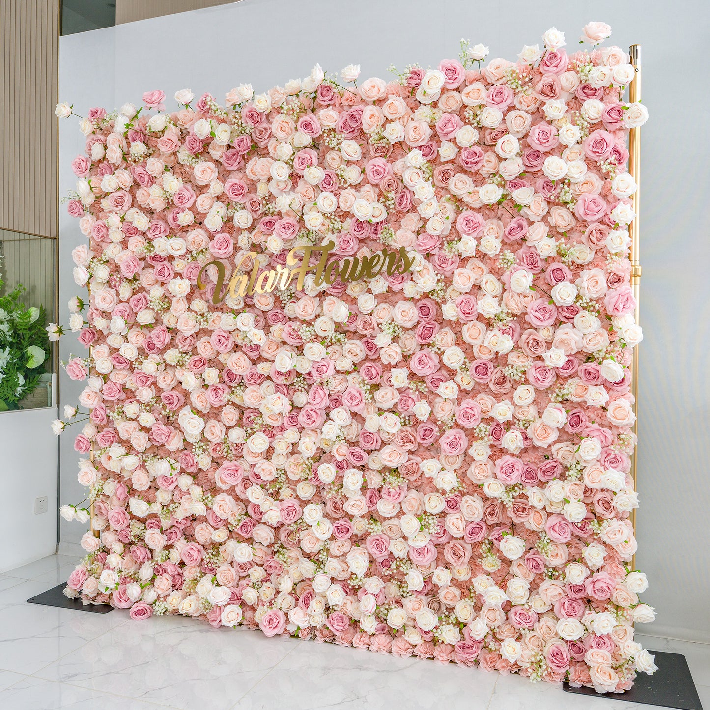 This photo showcases a large floral backdrop densely adorned with pink and white roses, interspersed with delicate baby's breath. The soft pastel color palette creates a romantic and elegant atmosphere. The words "Valar Flowers" in elegant gold lettering stand out prominently. Ideal for weddings or upscale events, it exudes a refined, dreamy style. 