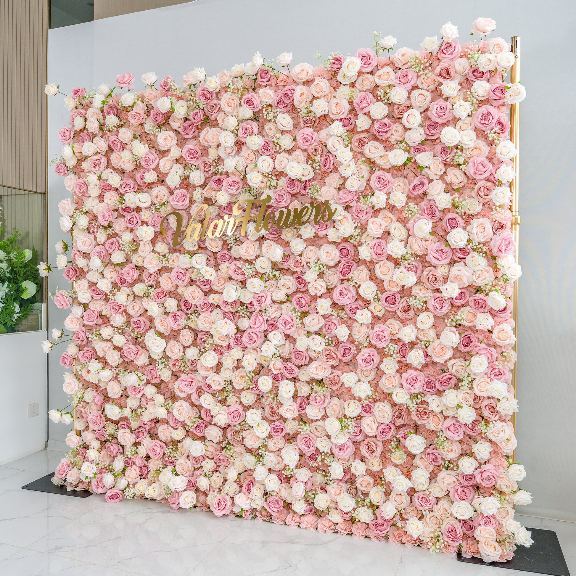 This photo showcases a large floral backdrop densely adorned with pink and white roses, interspersed with delicate baby's breath. The soft pastel color palette creates a romantic and elegant atmosphere. The words "Valar Flowers" in elegant gold lettering stand out prominently. Ideal for weddings or upscale events, it exudes a refined, dreamy style. 