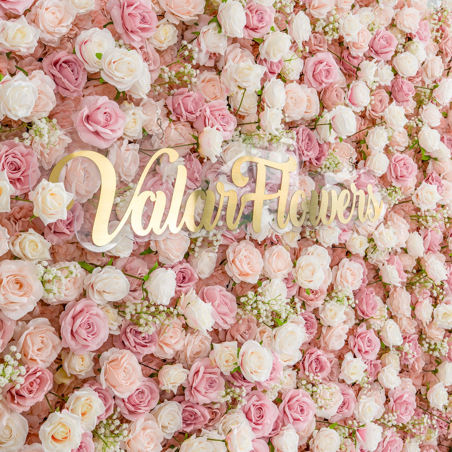 This photo showcases a lush floral backdrop composed of numerous pink and white roses, interspersed with delicate baby's breath. The soft pastel color scheme creates a romantic and dreamy atmosphere. Prominently displayed in elegant gold lettering is "Valar Flowers," adding a touch of sophistication. Ideal for weddings or romantic events, it exudes elegance and charm. 