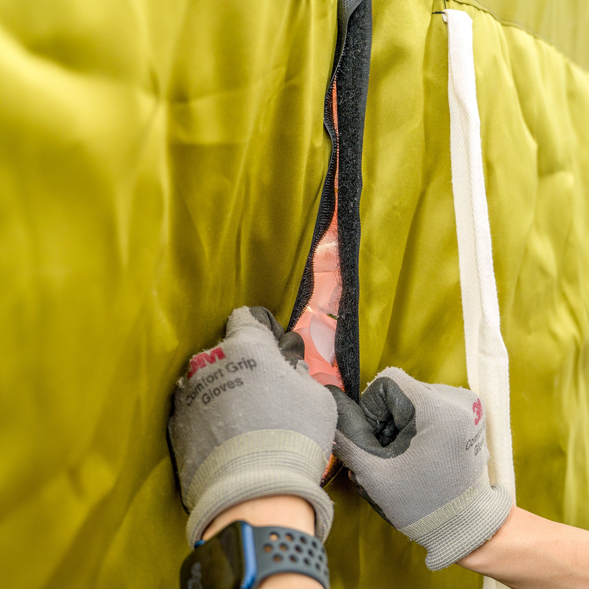 This photo shows a close - up of a pair of hands wearing grey "Comfort Grip Gloves." The hands are actively engaged in fastening or unfastening a black hook - and - loop fastener on a bright yellow fabric. The fabric appears to be part of a larger structure, possibly a tent or cover, with a white strap visible on the right side. The focus is on the action of securing the material, highlighting the gloves' functionality. 