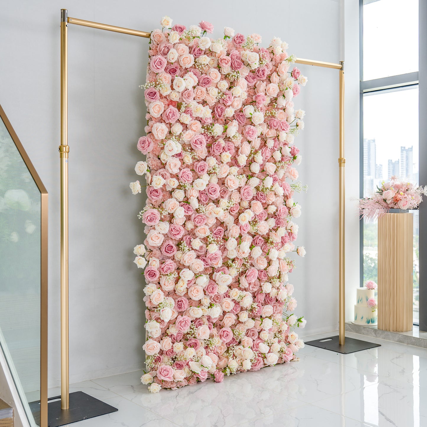 This photo showcases a large, elegant floral backdrop densely covered with pink and white roses. The backdrop is mounted on a golden metal frame, standing against a light - colored wall. The setting appears to be an indoor space with a marble - tiled floor and large windows, through which a cityscape is faintly visible. To the right, there's a decorative arrangement in a tall, wooden - textured vase, enhancing the overall romantic and sophisticated ambiance, ideal for events like weddings. 