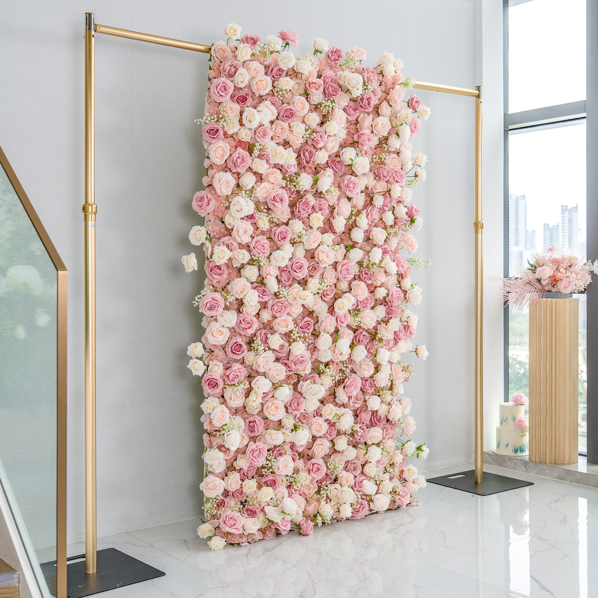 This photo showcases a large, elegant floral backdrop densely covered with pink and white roses. The backdrop is mounted on a golden metal frame, standing against a light - colored wall. The setting appears to be an indoor space with a marble - tiled floor and large windows, through which a cityscape is faintly visible. To the right, there's a decorative arrangement in a tall, wooden - textured vase, enhancing the overall romantic and sophisticated ambiance, ideal for events like weddings. 