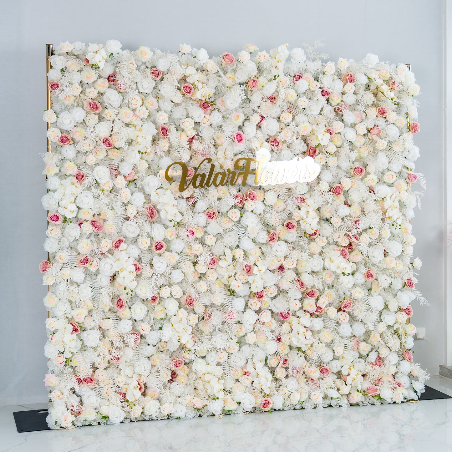 This photo features a lush floral backdrop densely covered with white roses, interspersed with delicate pink roses and some white foliage. The soft color palette of white and light pink creates a serene and elegant atmosphere. The words "Valar Flowers" in elegant gold lettering stand out prominently. Ideal for weddings, upscale events, or romantic occasions, it exudes a refined and dreamy style. 