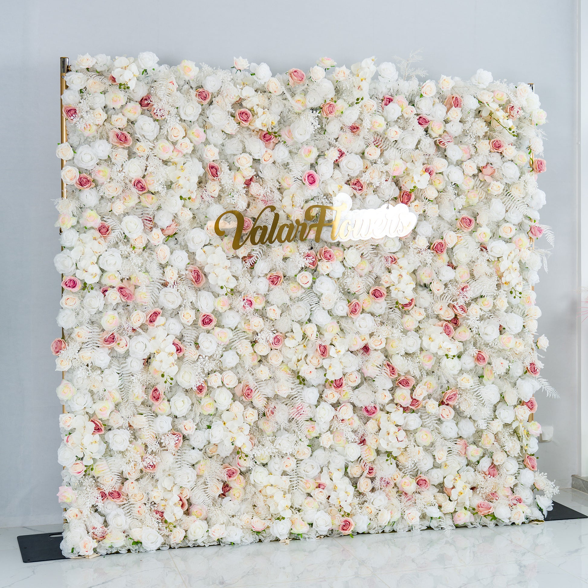 This photo features a lush floral backdrop densely covered with white roses, interspersed with delicate pink roses and some white foliage. The soft color palette of white and light pink creates a serene and elegant atmosphere. The words "Valar Flowers" in elegant gold lettering stand out prominently. Ideal for weddings, upscale events, or romantic occasions, it exudes a refined and dreamy style. 