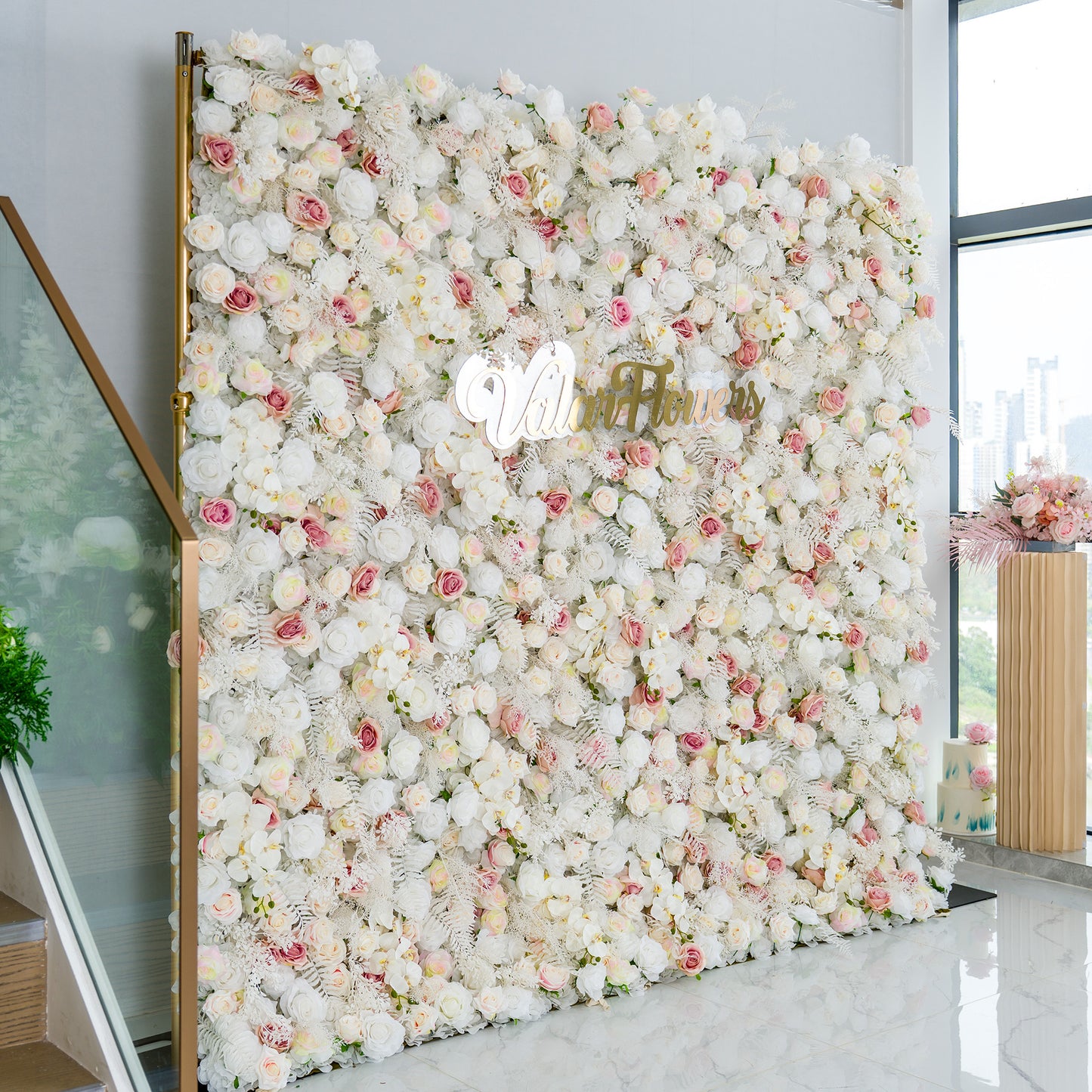 This photo showcases a large floral backdrop densely adorned with white roses, interspersed with delicate pink roses and some white foliage. The soft color palette of white and light pink creates a serene and elegant atmosphere. The words "Valar Flowers" in elegant gold lettering stand out prominently. Ideal for weddings, upscale galas, or sophisticated events, it exudes a refined and dreamy style. 