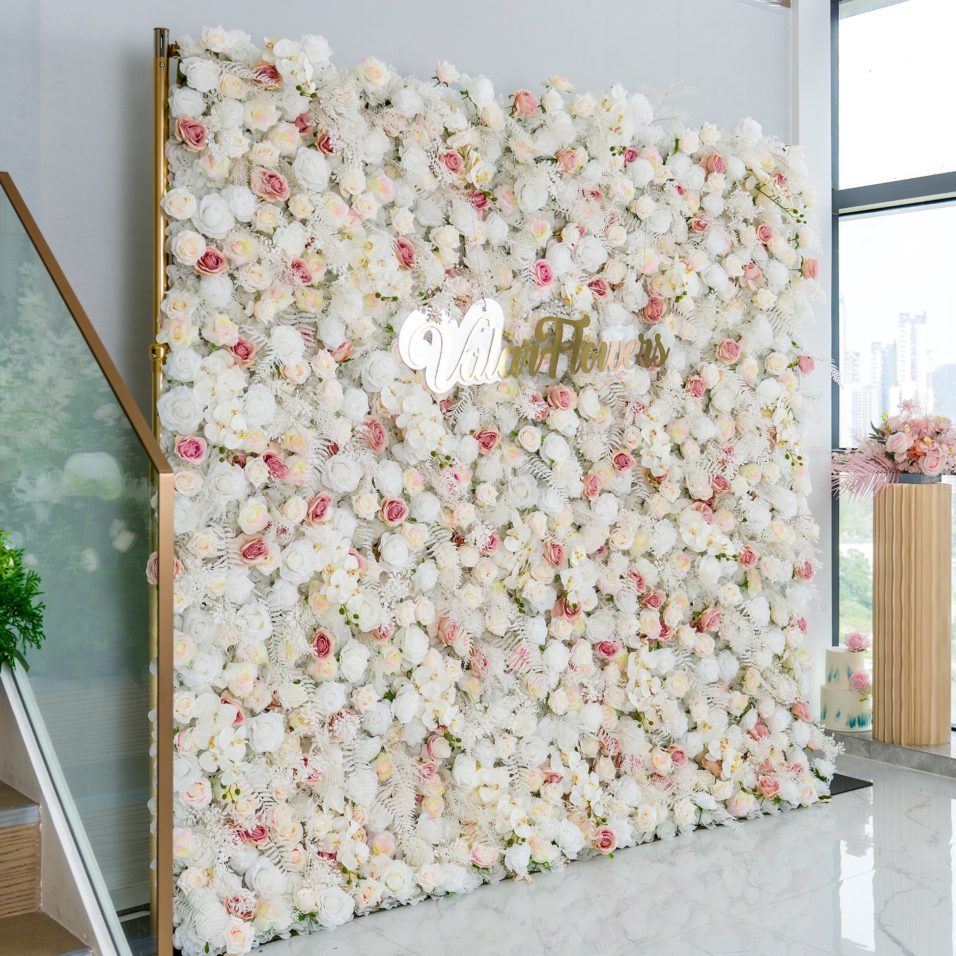 This photo showcases a large floral backdrop densely adorned with white roses, interspersed with delicate pink roses and some white foliage. The soft color palette of white and light pink creates a serene and elegant atmosphere. The words "Valar Flowers" in elegant gold lettering stand out prominently. Ideal for weddings, upscale galas, or sophisticated events, it exudes a refined and dreamy style. 