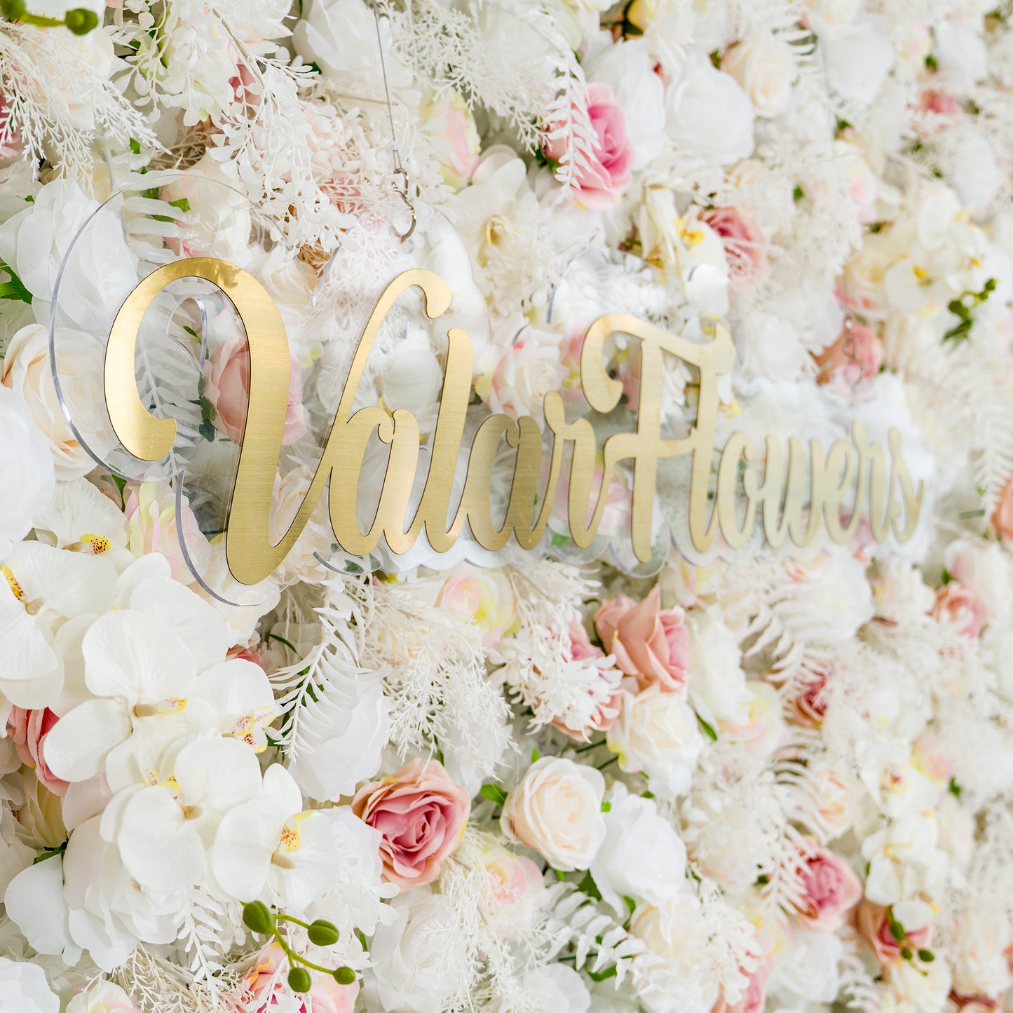 This photo showcases a lush floral backdrop filled with white roses, pink - tinged roses, and white orchids, complemented by delicate white foliage. The soft pastel color palette of white and light pink creates an elegant and romantic atmosphere. The words "Valar Flowers" in elegant gold lettering stand out prominently. Ideal for weddings or upscale events, it exudes a refined, dreamy style. 