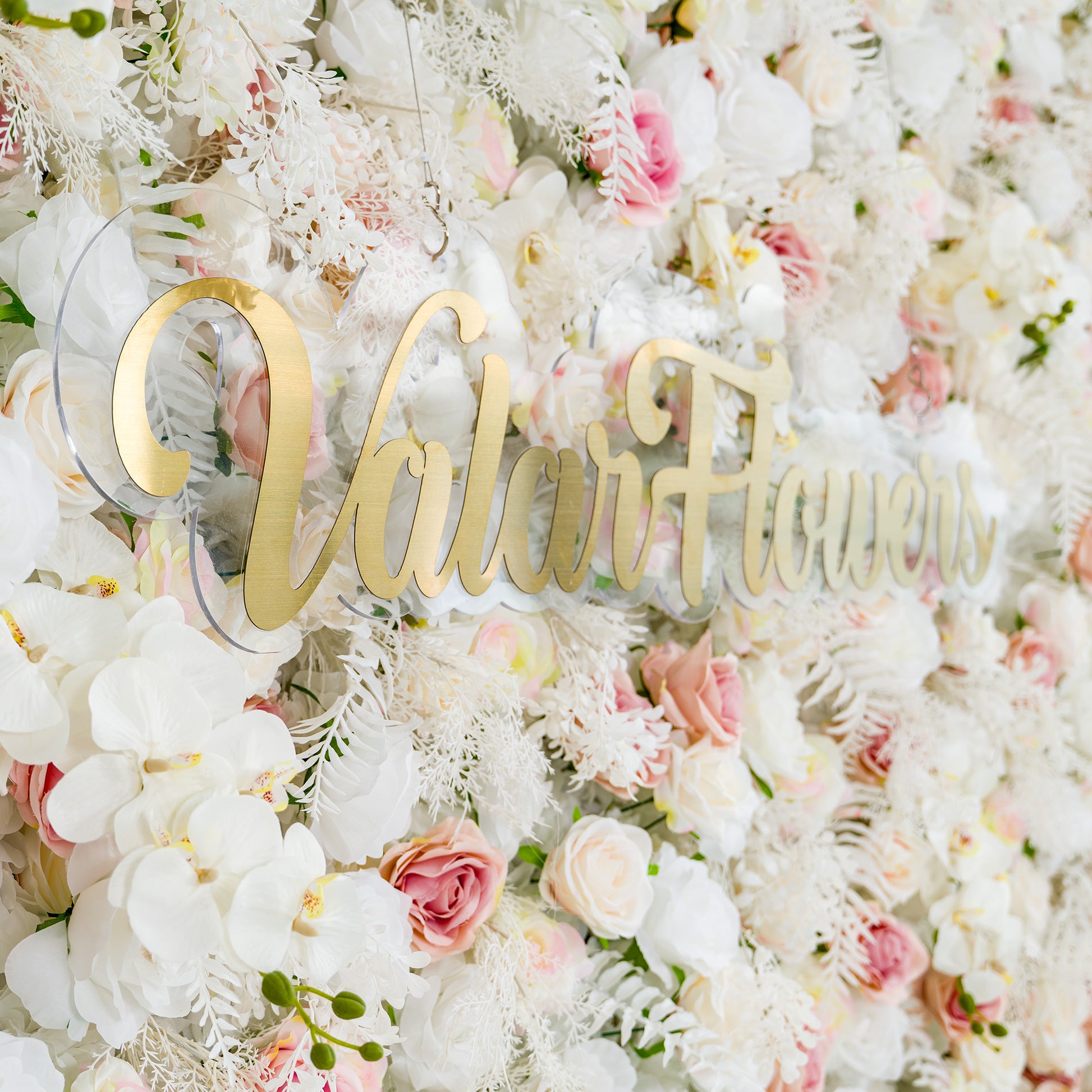 This photo showcases a lush floral backdrop filled with white roses, pink - tinged roses, and white orchids, complemented by delicate white foliage. The soft pastel color palette of white and light pink creates an elegant and romantic atmosphere. The words "Valar Flowers" in elegant gold lettering stand out prominently. Ideal for weddings or upscale events, it exudes a refined, dreamy style. 