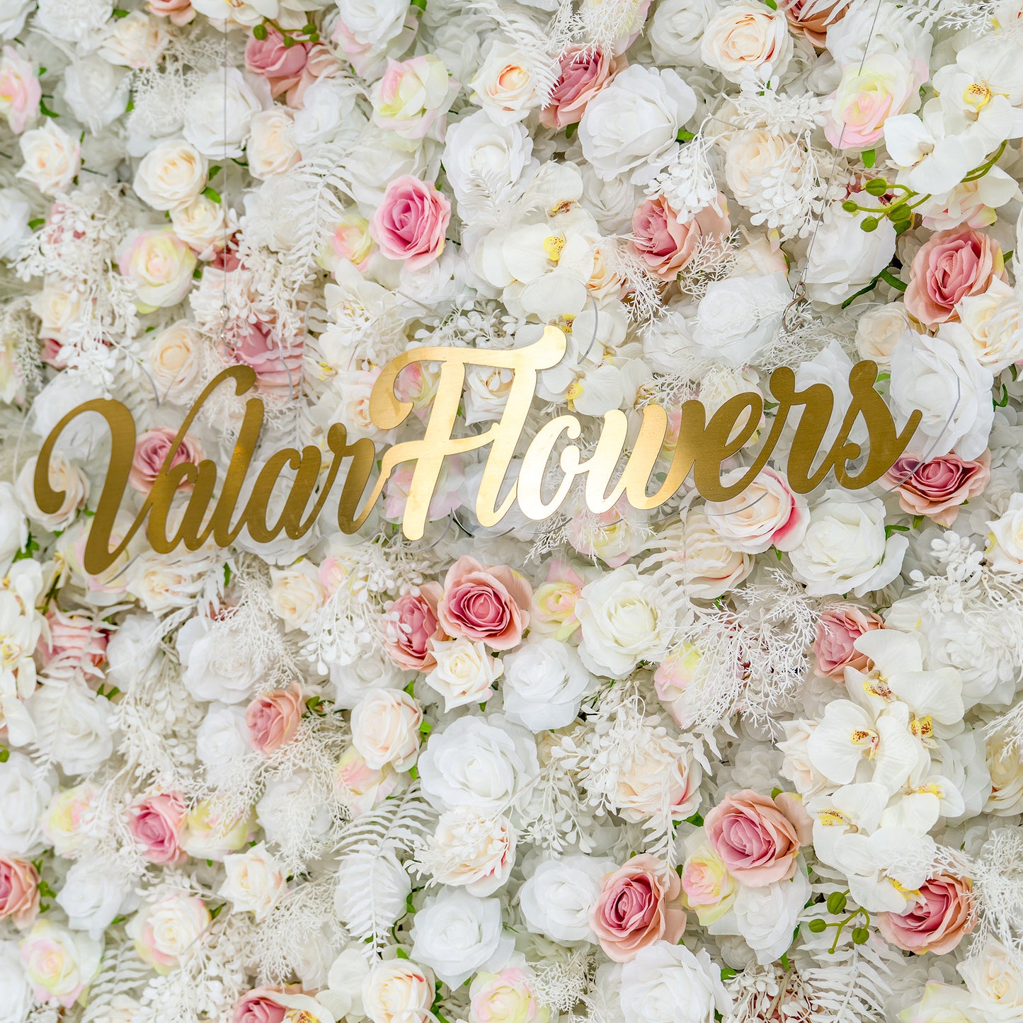 This photo showcases a lush floral backdrop with white roses, pink roses, and white orchids, complemented by white foliage. The soft color palette of white, light pink, and hints of pastel yellow creates an elegant and romantic atmosphere. The golden text "Valar Flowers" stands out prominently. Ideal for weddings or upscale events, it exudes a refined, dreamy style. 