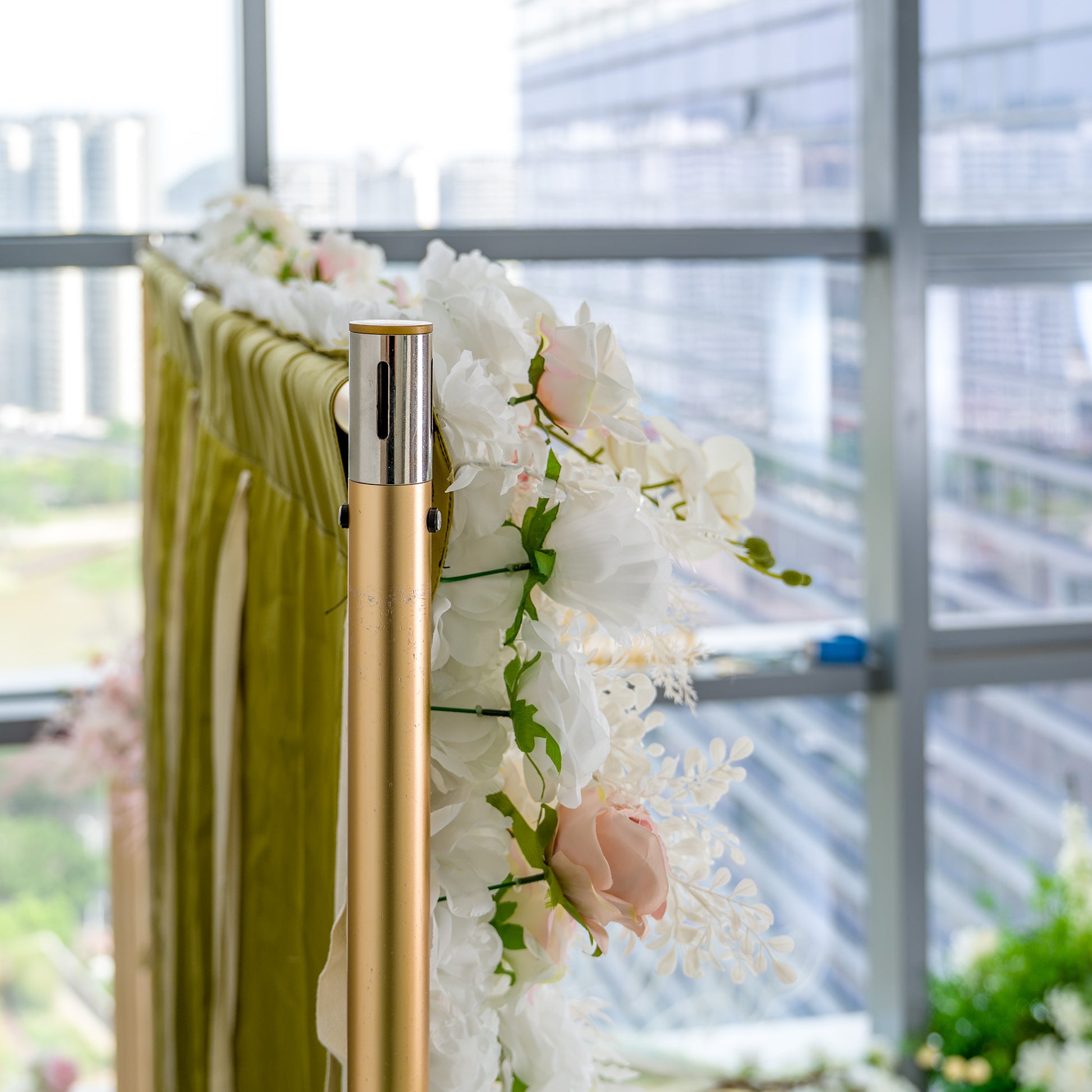This photo depicts a section of a floral - adorned structure. White roses and pink - tinted roses are prominently featured, complemented by green leaves and some white foliage. The flowers are arranged on a golden pole, with a backdrop of green draped fabric. Set against a cityscape view through large windows, it exudes an elegant and refined style, ideal for upscale events like weddings or galas. 