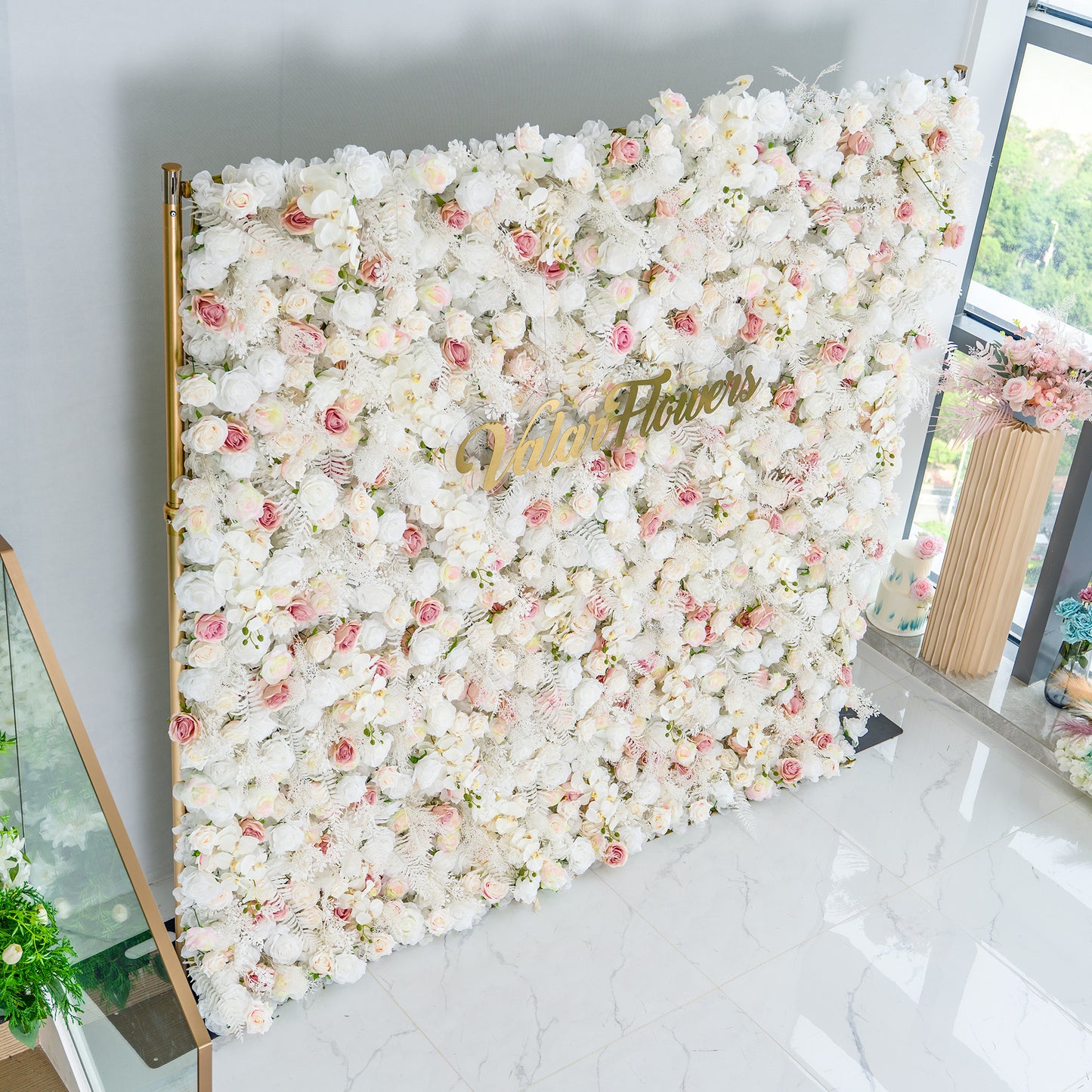 This photo showcases a large floral backdrop densely adorned with white roses, interspersed with delicate pink roses and white foliage. The soft color palette of white and light pink creates an elegant and serene atmosphere. The words "Valar Flowers" in elegant gold lettering stand out prominently. Ideal for weddings or upscale events, it exudes a refined, dreamy style. 