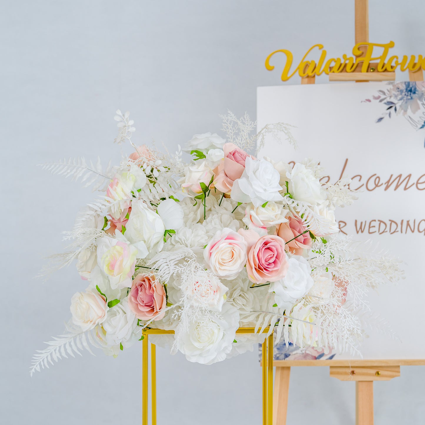 This photo showcases a lush floral arrangement, featuring a blend of white and pink roses, complemented by delicate white foliage. The flowers are artfully arranged in a golden - framed stand, exuding elegance. In the background, an easel displays a sign with "Welcome to our wedding" text and floral accents, enhancing the romantic ambiance. Ideal for weddings, it radiates a dreamy, refined style. 