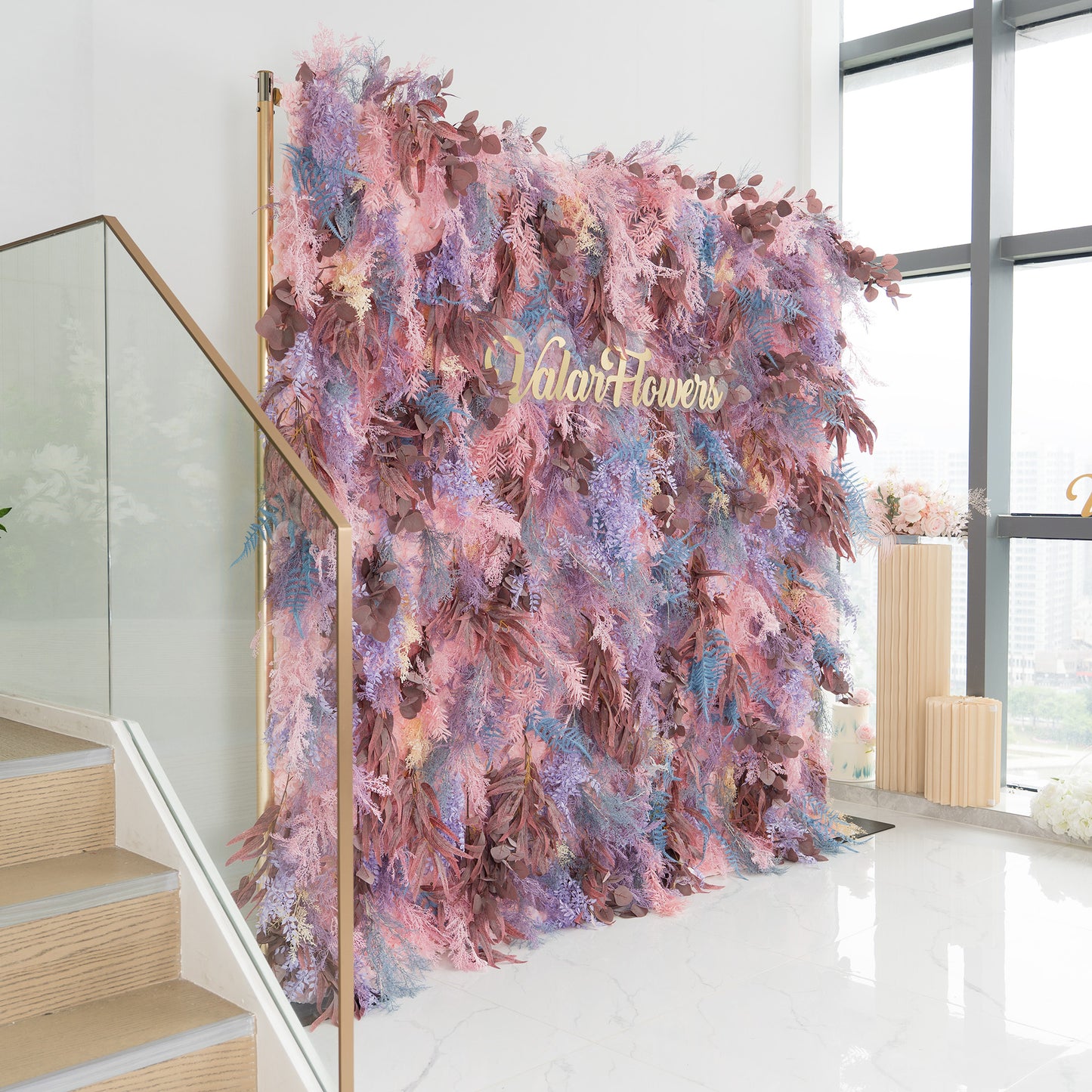 This backdrop is a lush, eye - catching installation. It's densely adorned with foliage in a dreamy color palette of pinks, purples, and blues, interspersed with rich brown leaves. The words "Valar Flowers" in elegant gold lettering stand out prominently. Situated near a staircase with a glass railing and beside large windows, it bathes in natural light. Ideal for events like weddings or upscale gatherings, it exudes a romantic and whimsical charm. 
