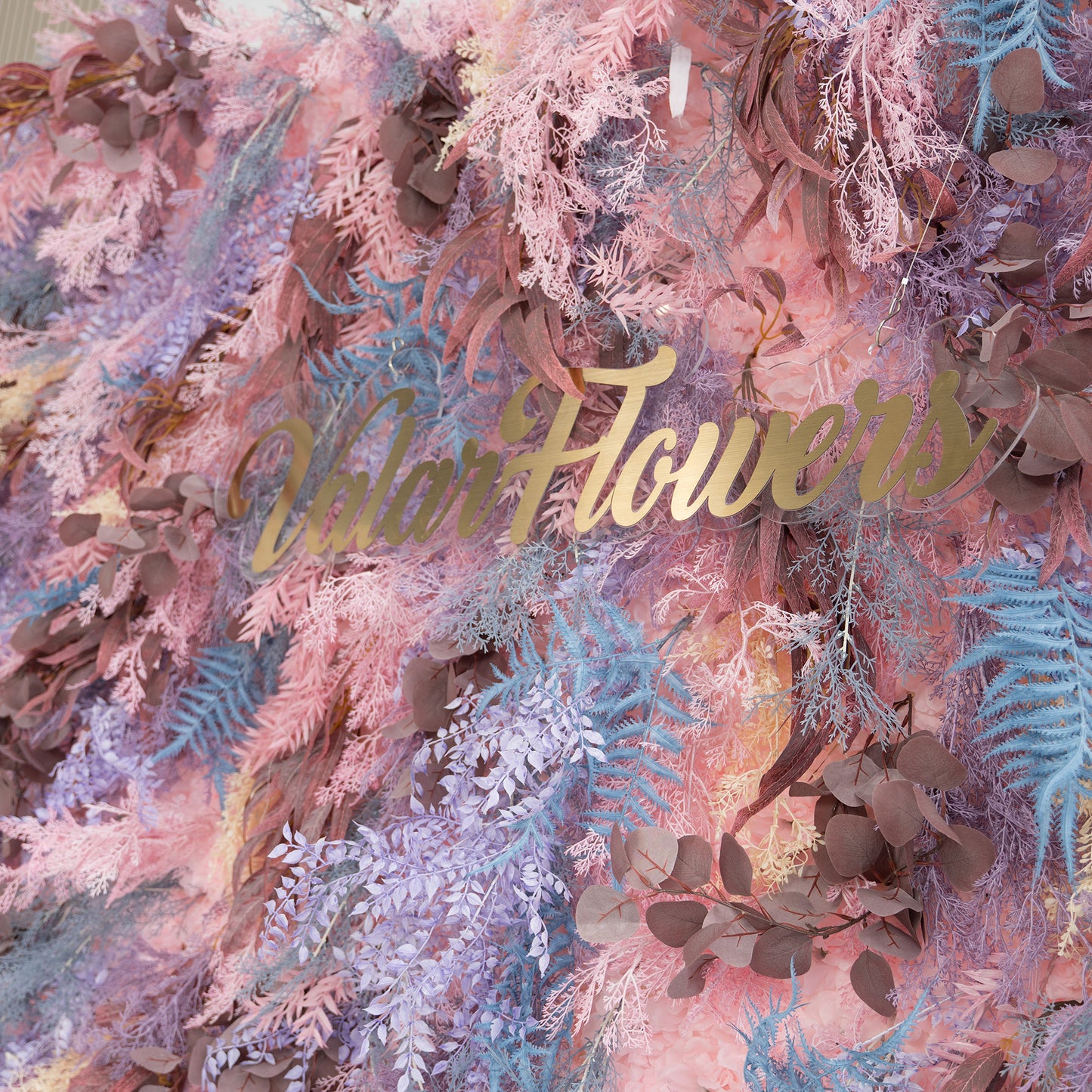 This photo showcases a lush foliage backdrop with a dreamy color scheme. It features a variety of plants, including fern - like fronds in shades of blue and purple, along with eucalyptus leaves in earthy brown tones, all interwoven with soft pink foliage. The "Valar Flowers" sign in elegant gold lettering stands out. Ideal for weddings, parties, or upscale events, it exudes a romantic and whimsical style. 