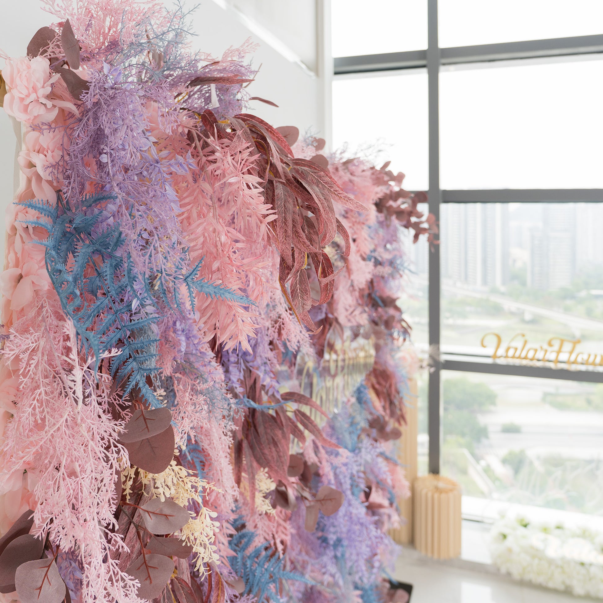 This photo showcases a lush foliage backdrop, a vibrant mix of plants. It features fern - like fronds in blue and purple, along with eucalyptus leaves in earthy browns, all interspersed with soft pink foliage. The elegant "Valar Flowers" sign in gold stands out. Ideal for weddings, parties, or upscale events, it exudes a romantic and whimsical style, enhanced by natural light from the nearby windows. 