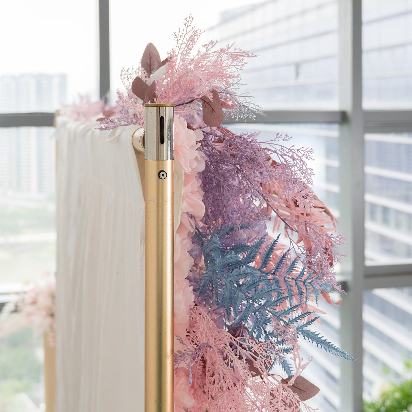 This photo captures a close - up of a decorative foliage backdrop. It features an assortment of plants with fern - like fronds in shades of blue and pink, along with eucalyptus - like leaves in earthy tones. The backdrop, attached to a golden pole, is set against a window with a cityscape view. Ideal for weddings, galas, or upscale parties, it exudes a romantic and elegant style with its pastel color palette. 