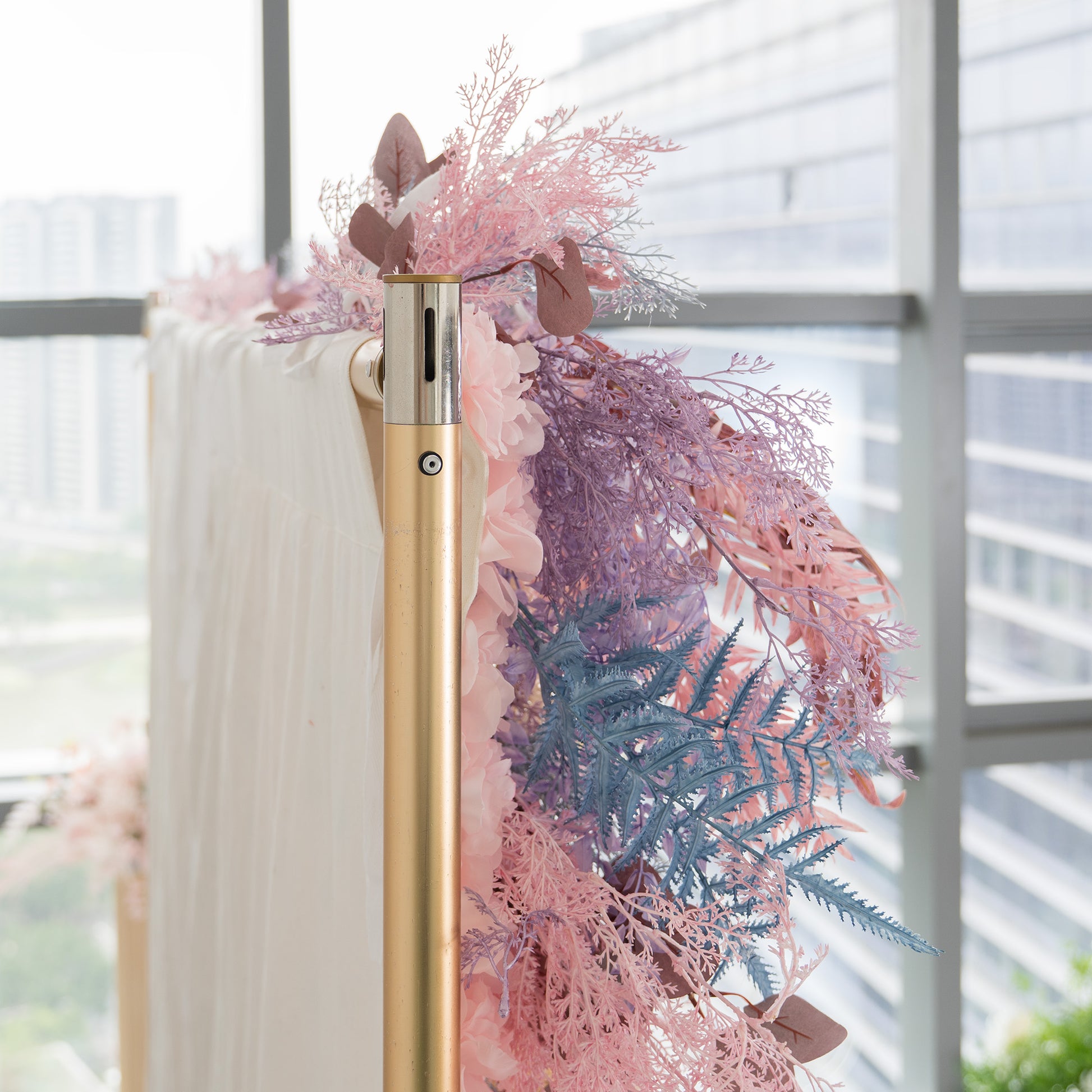 This photo captures a close - up of a decorative foliage backdrop. It features an assortment of plants with fern - like fronds in shades of blue and pink, along with eucalyptus - like leaves in earthy tones. The backdrop, attached to a golden pole, is set against a window with a cityscape view. Ideal for weddings, galas, or upscale parties, it exudes a romantic and elegant style with its pastel color palette. 