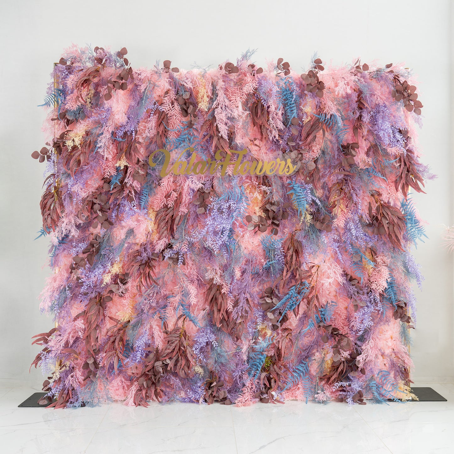 This photo showcases a lush foliage backdrop, a vibrant blend of plants. It features fern - like fronds in blue and purple, alongside eucalyptus - like leaves in earthy browns, all intertwined with soft pink foliage. The words "Valar Flowers" in elegant gold lettering stand out. Ideal for weddings, parties, or upscale events, it exudes a romantic and whimsical style. 