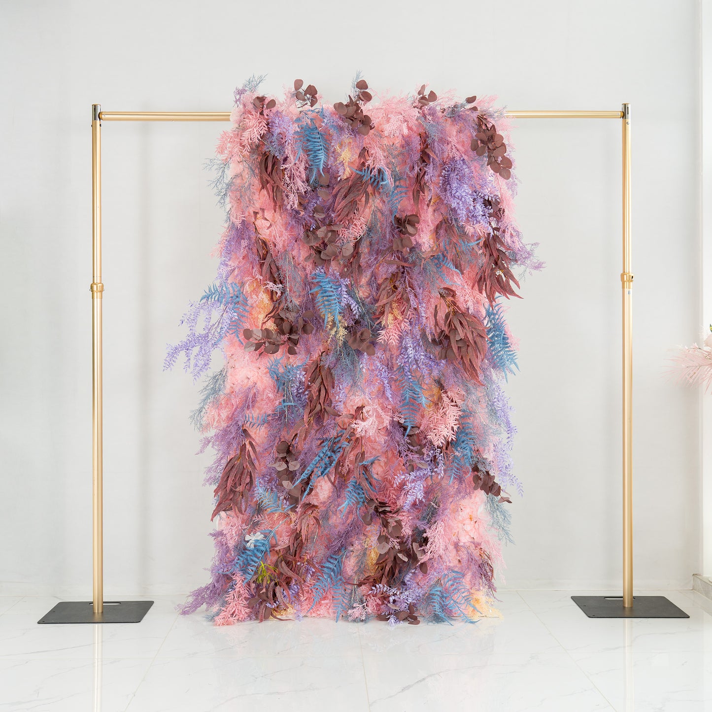This photo features a lush foliage backdrop supported by a golden frame. It combines fern - like fronds in shades of blue and purple with eucalyptus - like leaves in earthy brown tones, all interwoven with soft pink foliage. The pastel color palette creates a dreamy look. Ideal for weddings, parties, or upscale events, it exudes a romantic and whimsical style. 