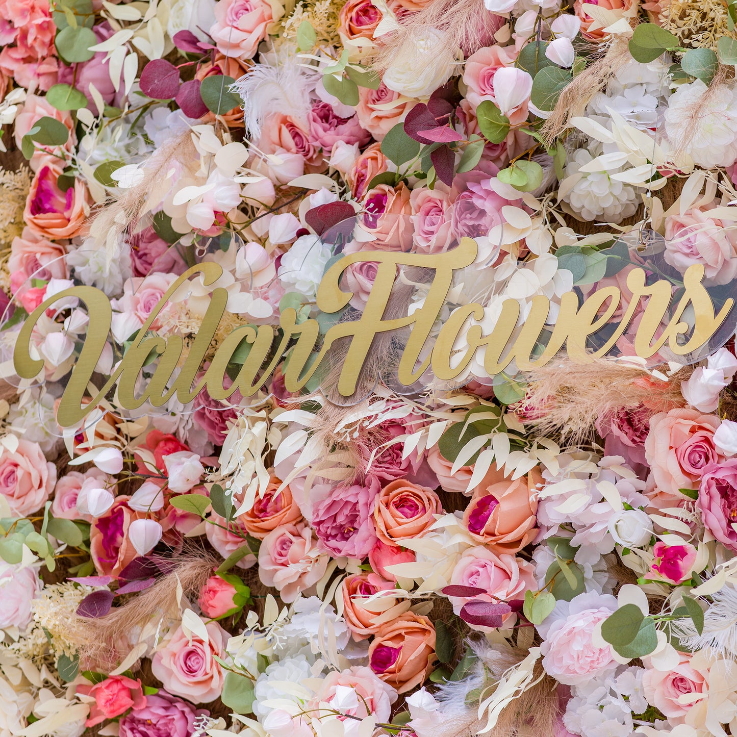 This photo showcases a lush, colorful floral backdrop densely packed with an array of artificial roses and peonies in soft pink, peach, and white hues. Interspersed with green eucalyptus leaves and delicate pampas grass, it exudes romance. The sign “ValarFlowers” in elegant gold lettering stands out. Ideal for weddings, bridal showers, or upscale events, it creates a dreamy, elegant atmosphere. 