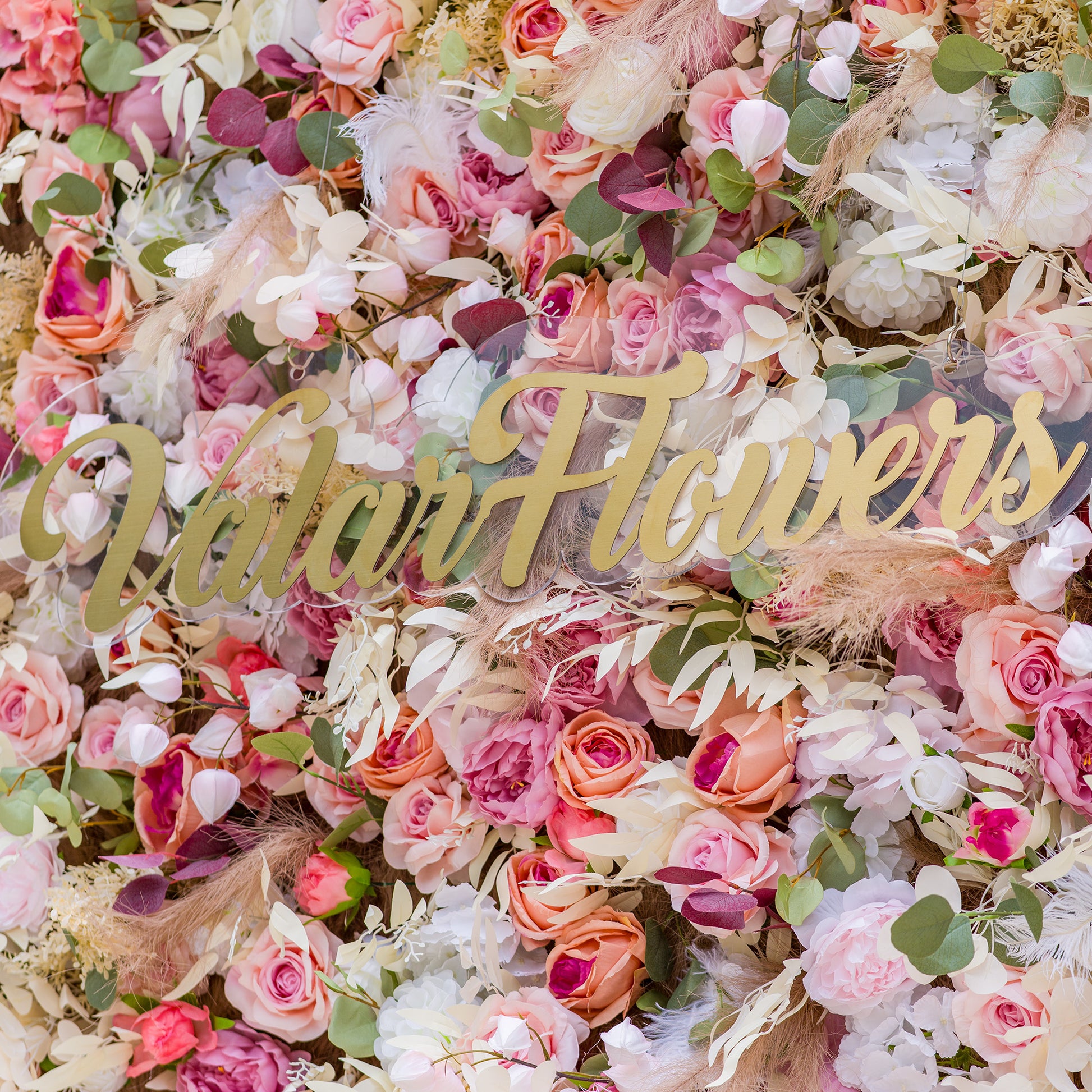 This photo showcases a lush, colorful floral backdrop densely packed with an array of artificial roses and peonies in soft pink, peach, and white hues. Interspersed with green eucalyptus leaves and delicate pampas grass, it exudes romance. The sign “ValarFlowers” in elegant gold lettering stands out. Ideal for weddings, bridal showers, or upscale events, it creates a dreamy, elegant atmosphere. 