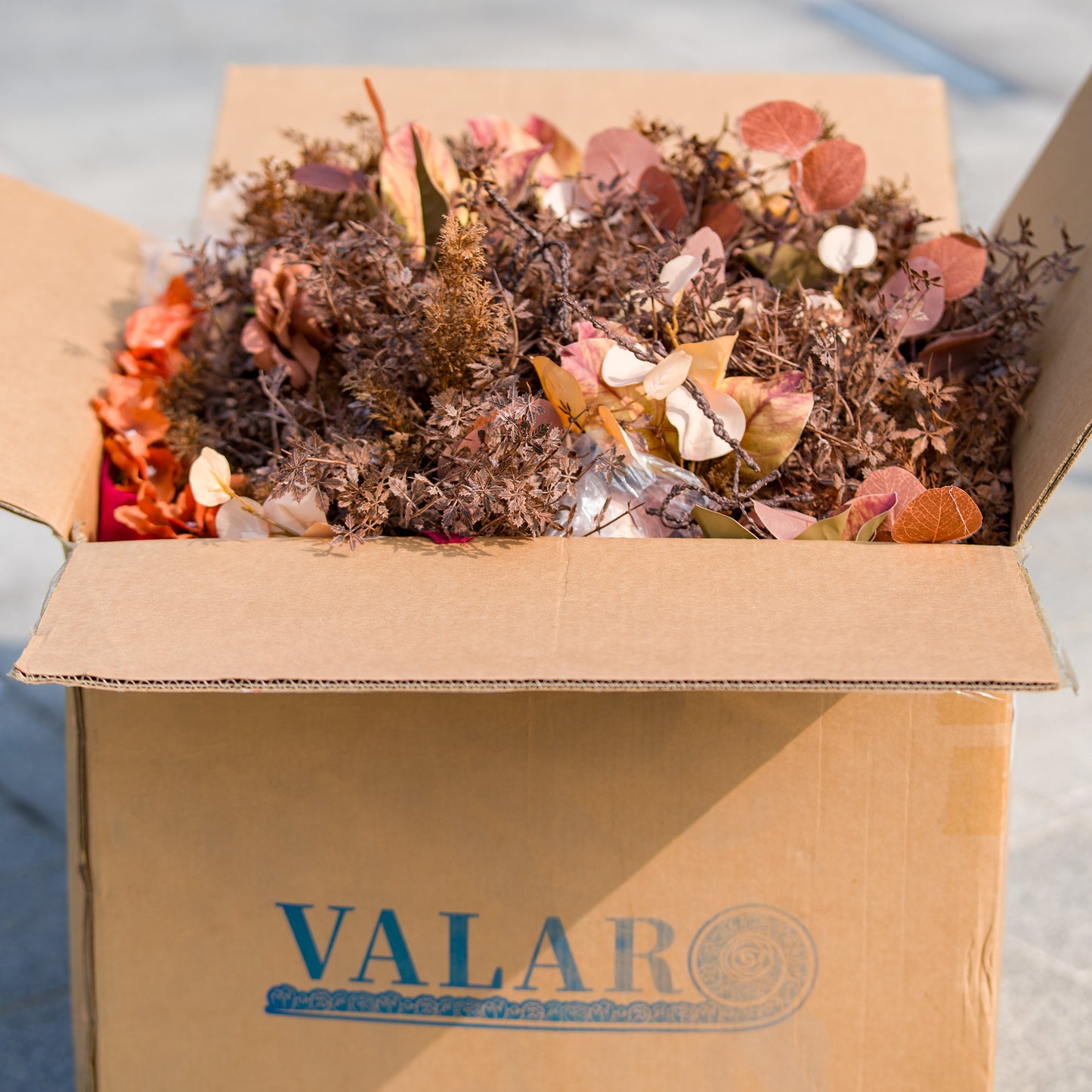 This photo shows an open cardboard box branded with “VALAR” filled with an assortment of autumn - colored artificial flowers and foliage in shades of rust, brown, and copper. Ideal for DIY event decor projects like creating a fall - themed backdrop for weddings or parties, it offers the materials to infuse seasonal charm. 
