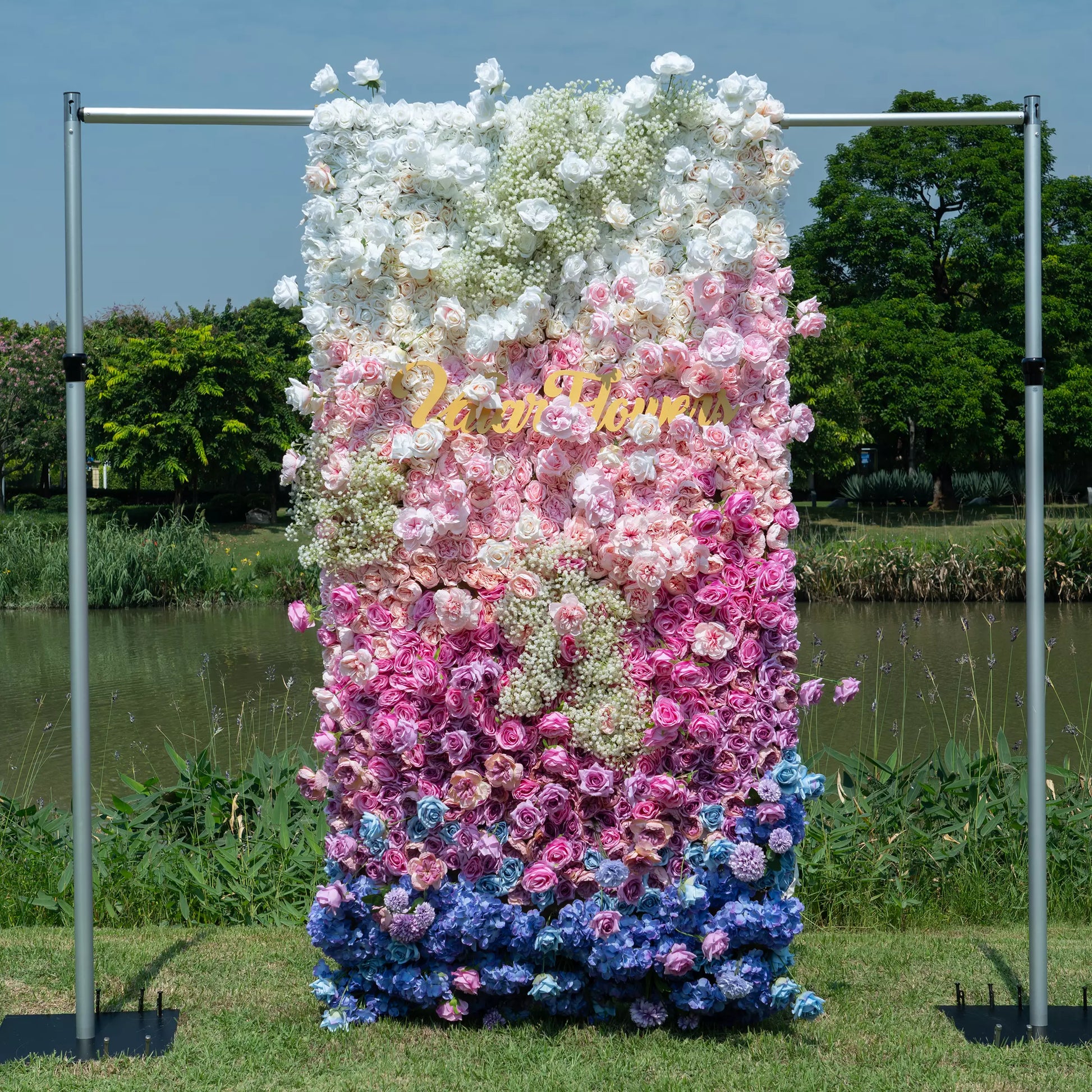 This photo features a stunning floral wall with a gradient of white, pink, purple, and blue flowers, including roses and hydrangeas, with baby's breath adding texture. "Valar Flowers" is displayed in elegant gold lettering. Set outdoors by a body of water with greenery, it has a dreamy, romantic style, perfect for weddings or photo - ops. 