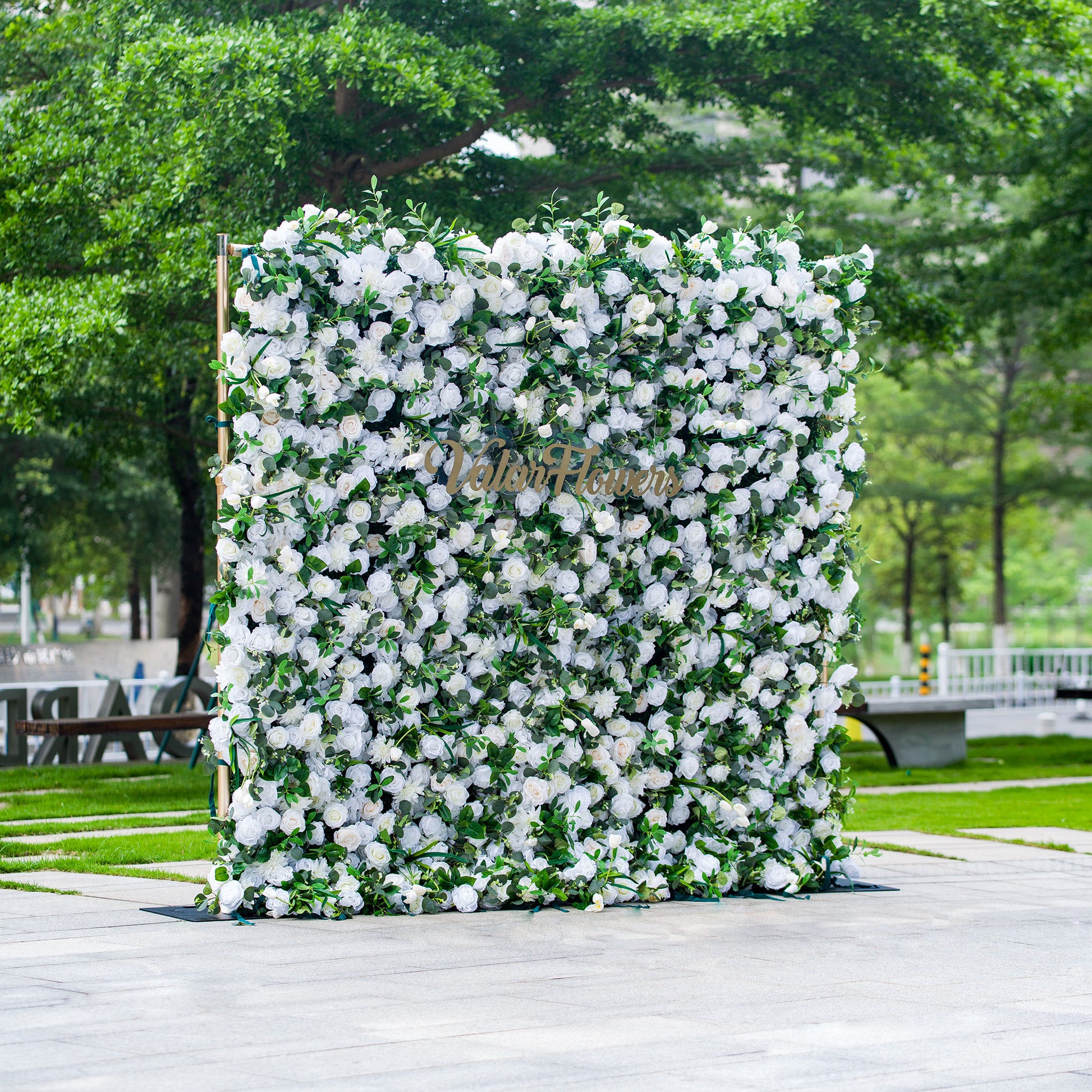 This photo features a large, square floral backdrop densely covered with white flowers, likely roses, interspersed with lush green foliage. The text “ValarFlowers” in elegant golden lettering stands out prominently. Ideal for events like a “White - Tie Garden Soirée” or a classic wedding, it exudes a sense of refined elegance, perfect for sophisticated outdoor celebrations. 