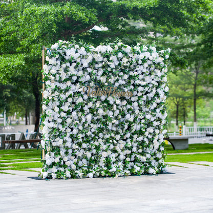 This photo features a large, square floral backdrop densely covered with white flowers, likely roses, interspersed with lush green foliage. The text “ValarFlowers” in elegant golden lettering stands out prominently. Ideal for events like a “White - Tie Garden Soirée” or a classic wedding, it exudes a sense of refined elegance, perfect for sophisticated outdoor celebrations. 
