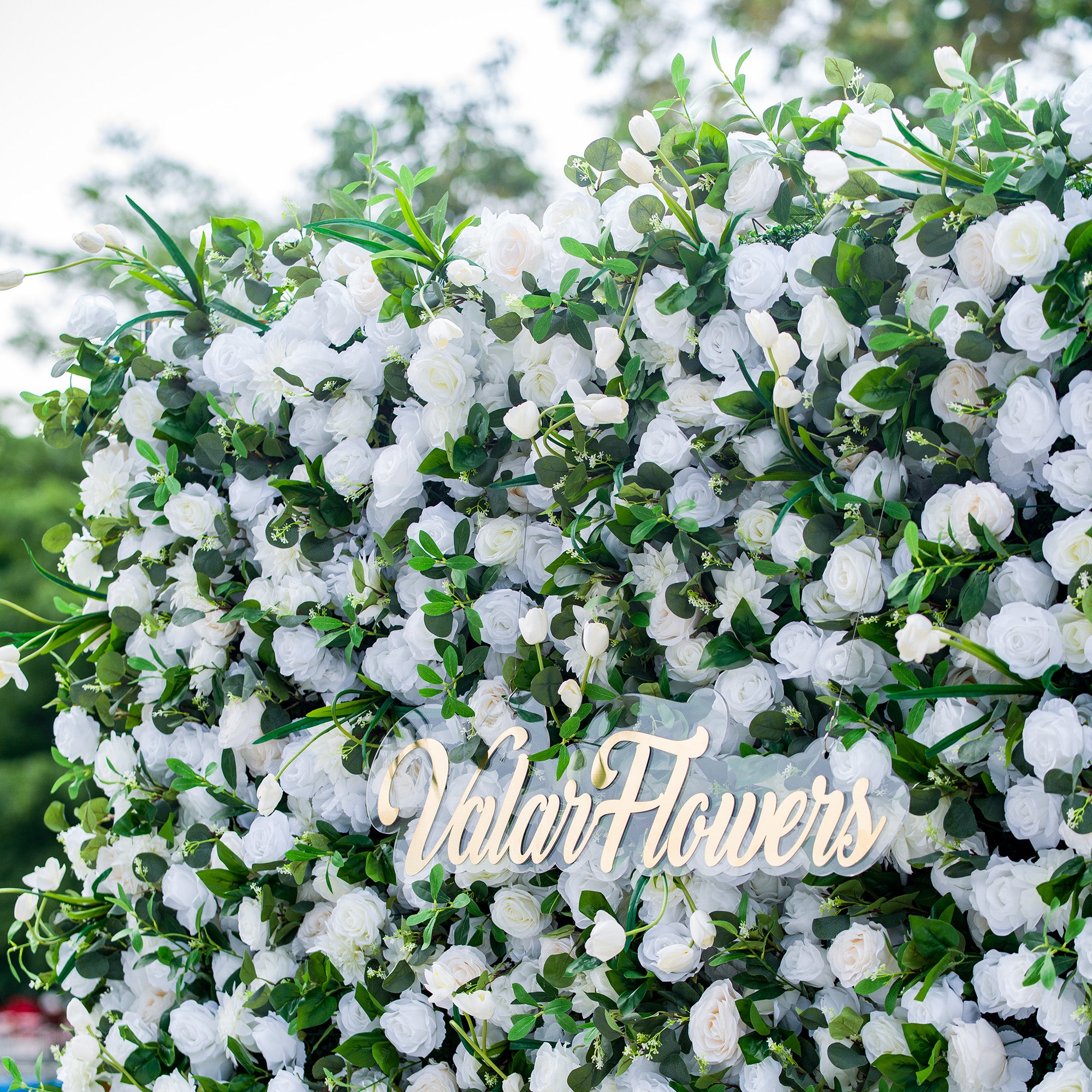 This photo showcases a lush floral backdrop densely filled with white roses, tulips, and other white blooms, interspersed with green foliage. The text “ValarFlowers” in elegant golden lettering stands out prominently. Ideal for events like a “Snow - White Soirée” or a refined garden wedding, it exudes a sense of purity and elegance, perfect for upscale and sophisticated occasions. 