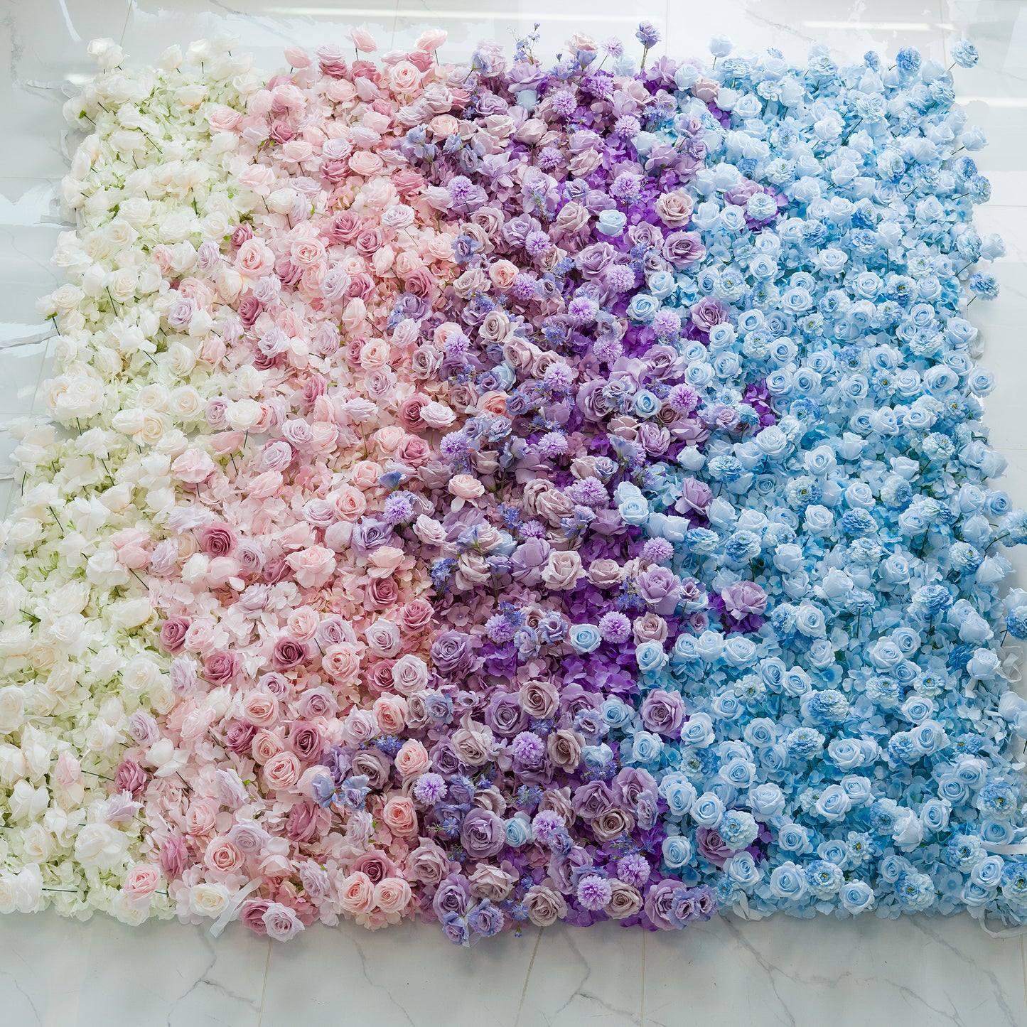 This photo features a stunning floral wall filled with roses in a gradient of colors—white, pink, purple, and blue. The ombre color theme creates a dreamy, elegant vibe, perfect for weddings, galas, or upscale events. The seamless blend of hues and abundant rose blooms form a sophisticated, photo - worthy backdrop, ideal for capturing memorable moments at celebrations seeking a touch of natural beauty and refined charm. 
