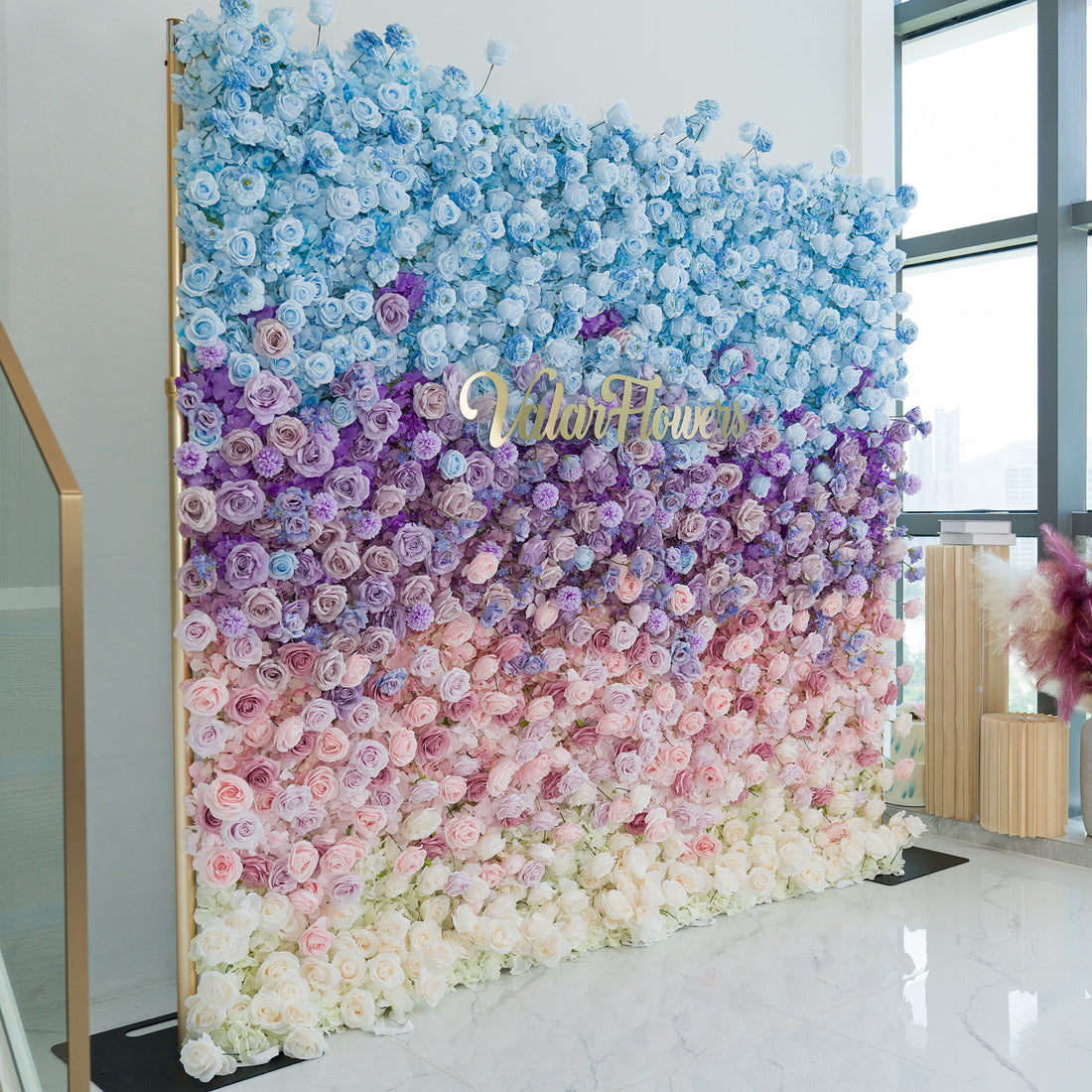 This striking floral wall features a gradient of roses, hydrangeas, and filler blooms in shades of blue, purple, pink, and white. The ombre color theme, transitioning smoothly from sky - blue at the top to soft white at the base, creates a dreamy, romantic vibe. A gold “ValarFlowers” sign adds elegance. Perfect for weddings, galas, or brand events, it serves as a sophisticated, photo - worthy backdrop, blending natural beauty with polished design to elevate any occasion. 