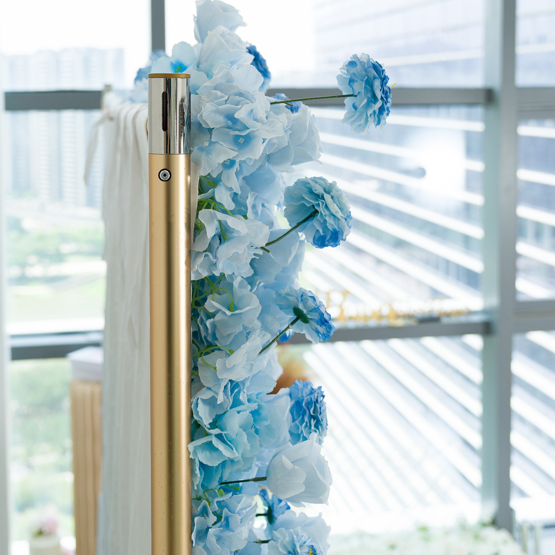 This photo features a gold pole adorned with blue hydrangeas and delicate blue flowers. The monochromatic light - blue color theme creates a serene, elegant vibe, perfect for events like weddings, baby showers, or upscale gatherings. The modern backdrop of glass windows and an urban skyline adds a sophisticated touch. The cohesive hue and realistic faux flowers offer a polished, long - lasting decorative element, ideal for setting a tranquil and refined mood at any celebration. 