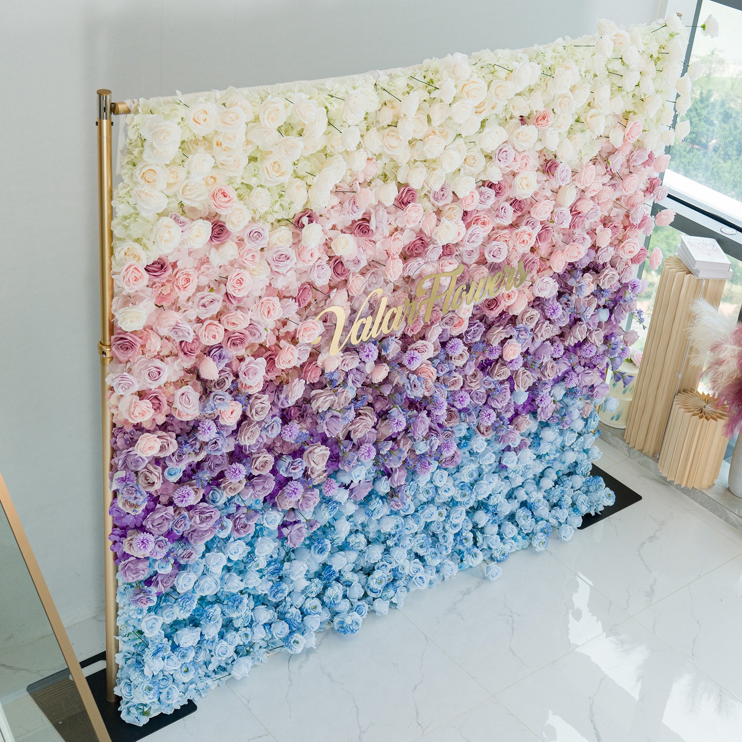 A captivating floral wall by ValarFlowers showcases a gradient of blooms—white roses at the top, transitioning through soft pink, lavender, to blue flowers. The gold “ValarFlowers” logo adds elegance. Set against a minimalist backdrop with a gold frame, this lush, colorful display is perfect for events like weddings or photo shoots, blending romance and sophistication. 