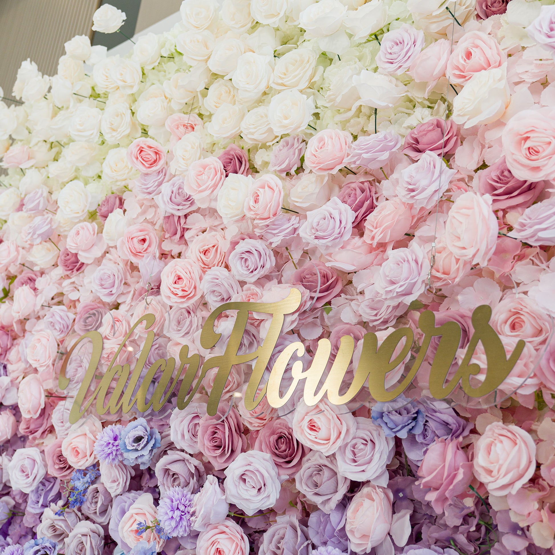 A lush floral wall features white roses, soft pink/lavender roses, and delicate filler flowers. The gold “ValarFlowers” sign adds elegance. The color theme—pastel gradients from white to purple—creates a romantic, dreamy vibe, perfect for weddings, events, or brand activations, blending natural beauty with polished charm. 