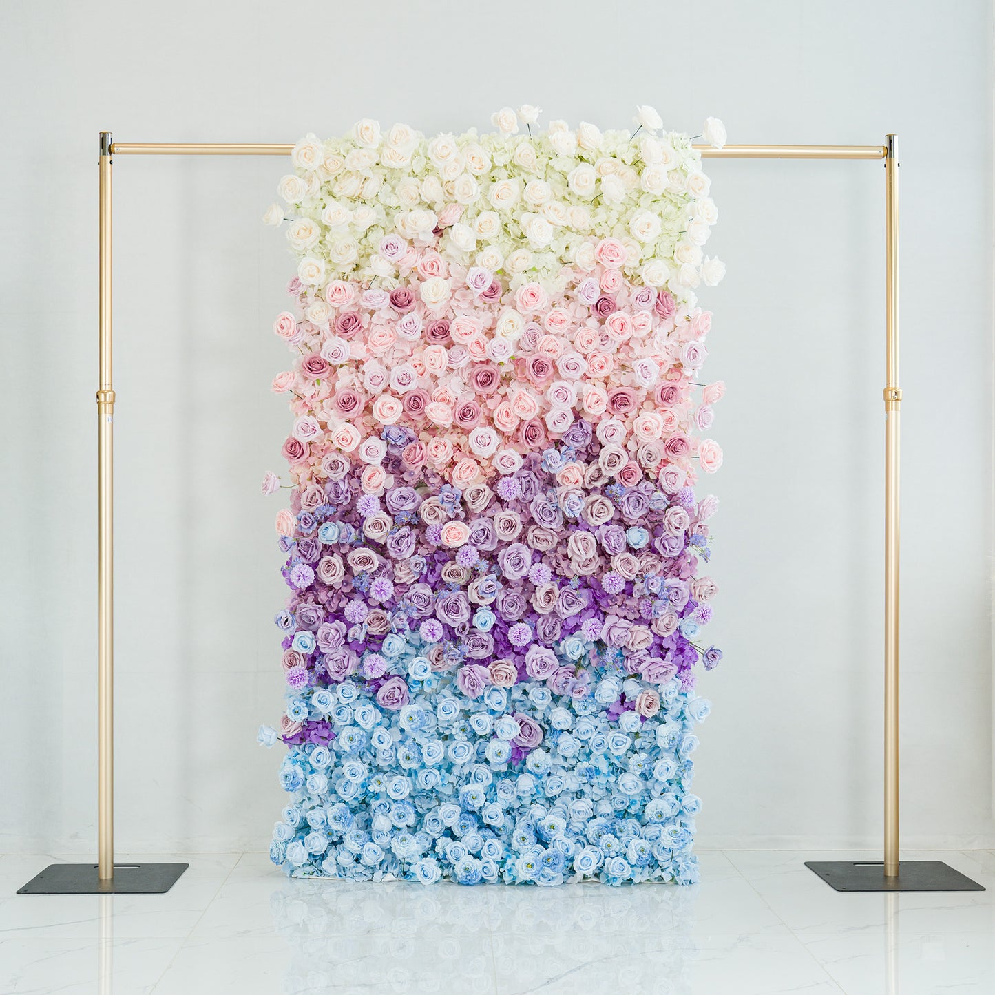 A 1.2x2.4m floral wall features a gradient of roses—white at the top, transitioning through pink, purple, to blue. Framed by gold poles, it’s set against a minimalist backdrop. The soft color theme creates an elegant, dreamy vibe, perfect for weddings, photo shoots, or upscale events, blending natural beauty with sophisticated design. 