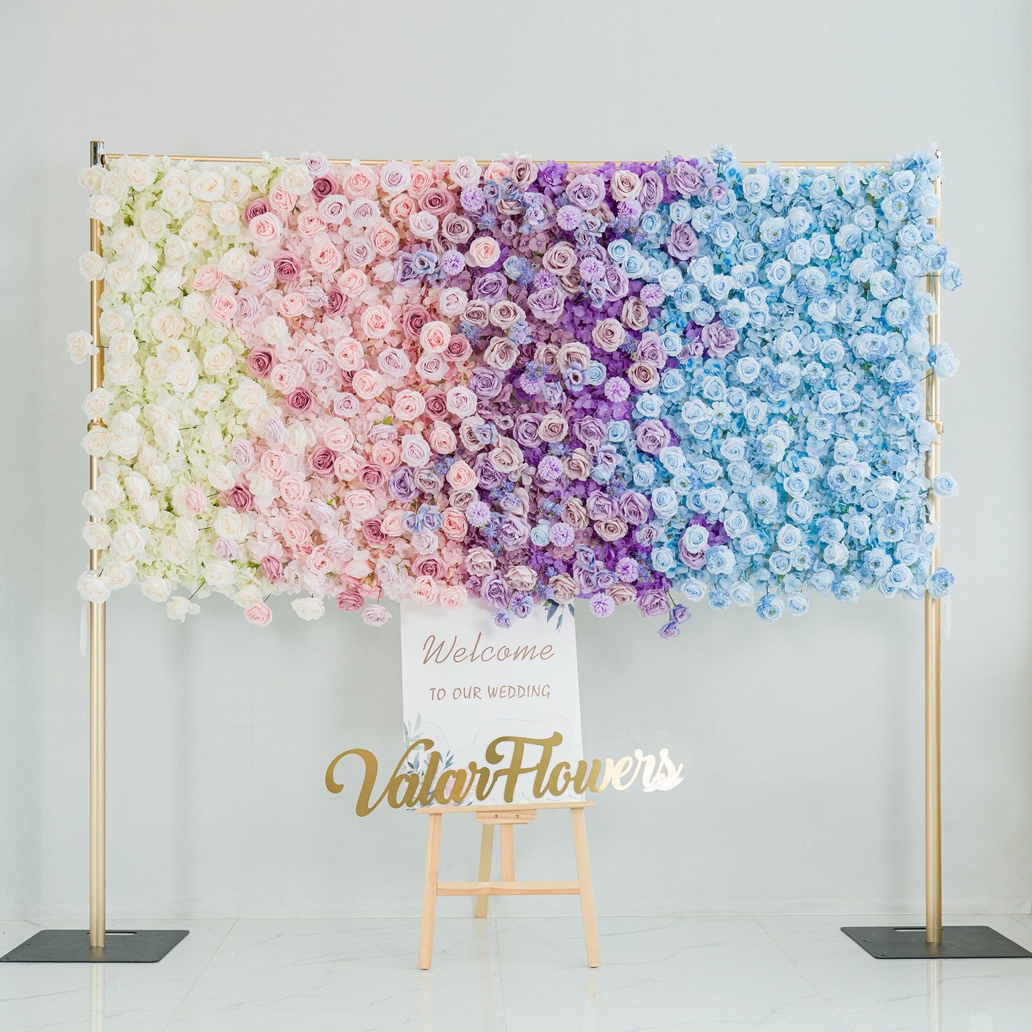 A stunning wedding - themed floral wall! It features a gradient of roses in white, soft pink, lavender, and blue. Baby’s breath and filler flowers add texture. The color theme—pastel to deeper hues—creates a romantic, dreamy vibe. A “Welcome to our wedding” sign and “ValarFlowers” logo complete the elegant, celebratory look, perfect for a wedding backdrop. 