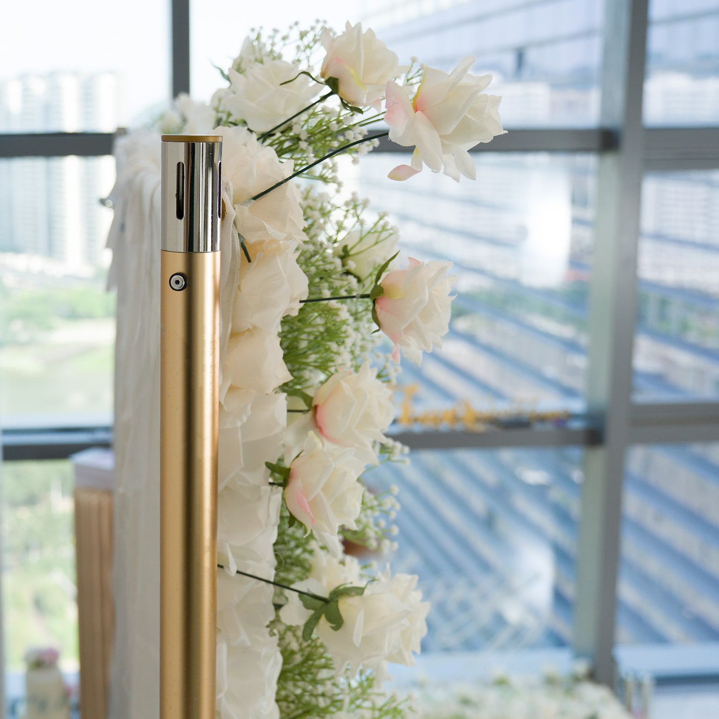A gold - toned pole adorned with a cascade of white roses and delicate baby’s breath, paired with flowing white fabric. Set against a backdrop of modern glass windows showing an urban scene, this elegant floral arrangement exudes a romantic, sophisticated vibe—perfect for weddings or upscale events, blending natural beauty with contemporary style. 