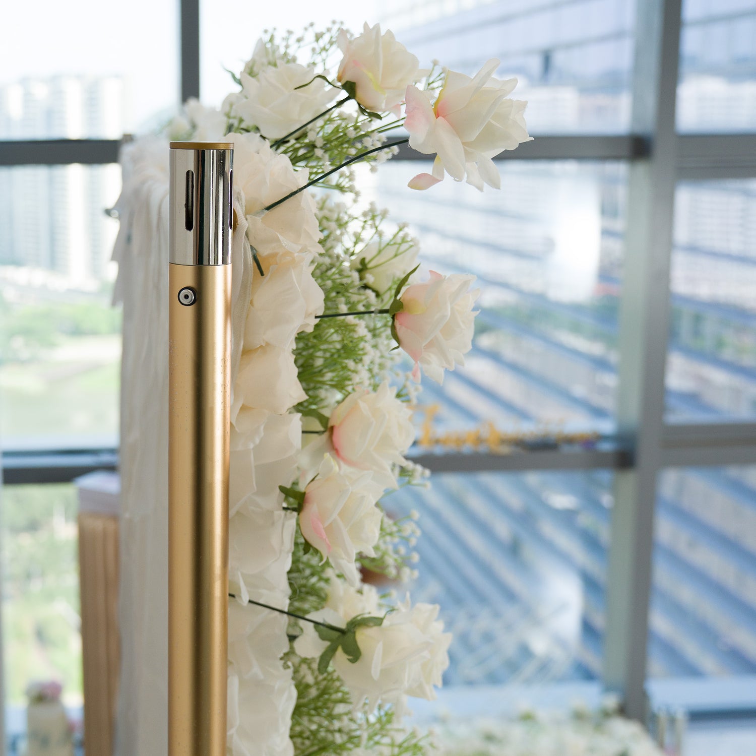 A gold - toned pole adorned with a cascade of white roses and delicate baby’s breath, paired with flowing white fabric. Set against a backdrop of modern glass windows showing an urban scene, this elegant floral arrangement exudes a romantic, sophisticated vibe—perfect for weddings or upscale events, blending natural beauty with contemporary style. 