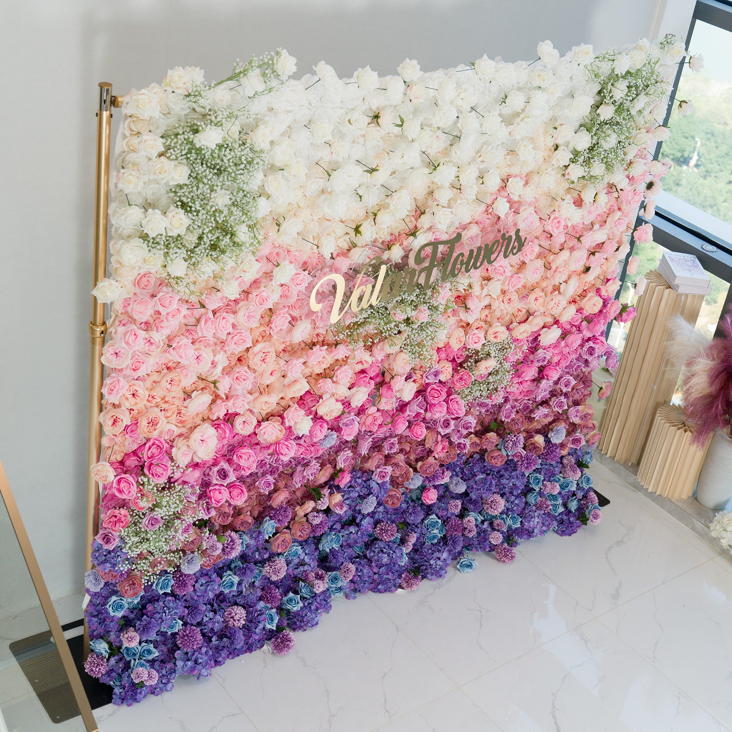This stunning floral wall by ValarFlowers features a gradient of blooms—white roses and baby’s breath at the top, transitioning to soft pink, lavender, and deep purple flowers. The gold “ValarFlowers” logo adds elegance. Set indoors with a marble floor and modern decor, it’s perfect for weddings, events, or photo backdrops, blending romance and sophistication. 