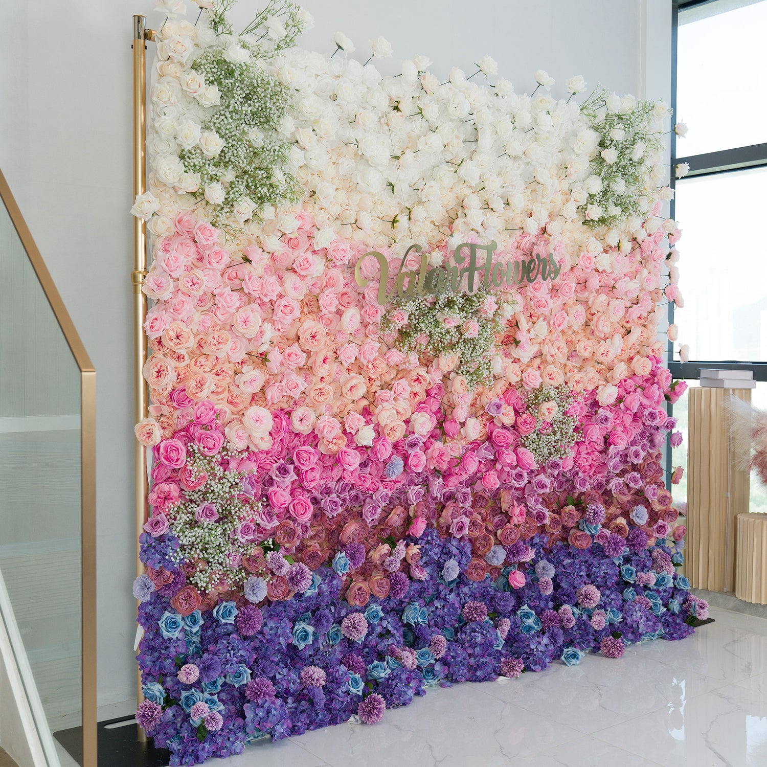 This stunning floral wall by ValarFlowers features a gradient of blooms—white roses and baby’s breath at the top, transitioning to soft pink, lavender, and deep purple flowers. The gold “ValarFlowers” logo adds elegance. Set indoors with a marble floor and modern decor, it’s perfect for weddings, events, or photo backdrops, blending romance and sophistication. 