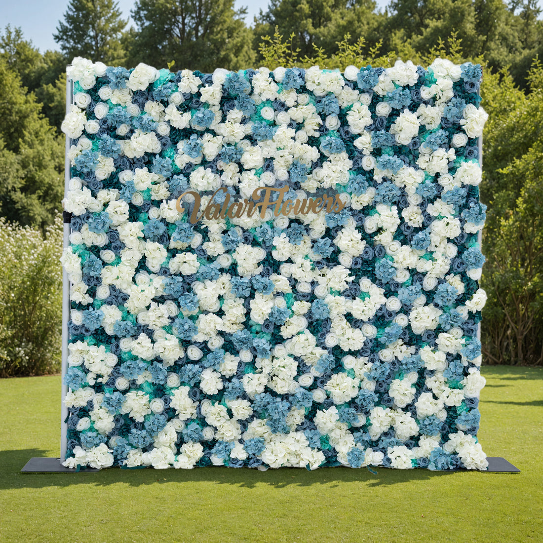 This photo features a stunning floral backdrop by Valar Flowers, showcasing a dense arrangement of artificial roses in white and varying shades of blue. The backdrop is set against a lush green lawn with trees in the background, creating a serene outdoor setting. The gold lettering of &quot;Valar Flowers&quot; adds a touch of elegance, making it an ideal decorative piece for weddings, photo shoots, or other special events.