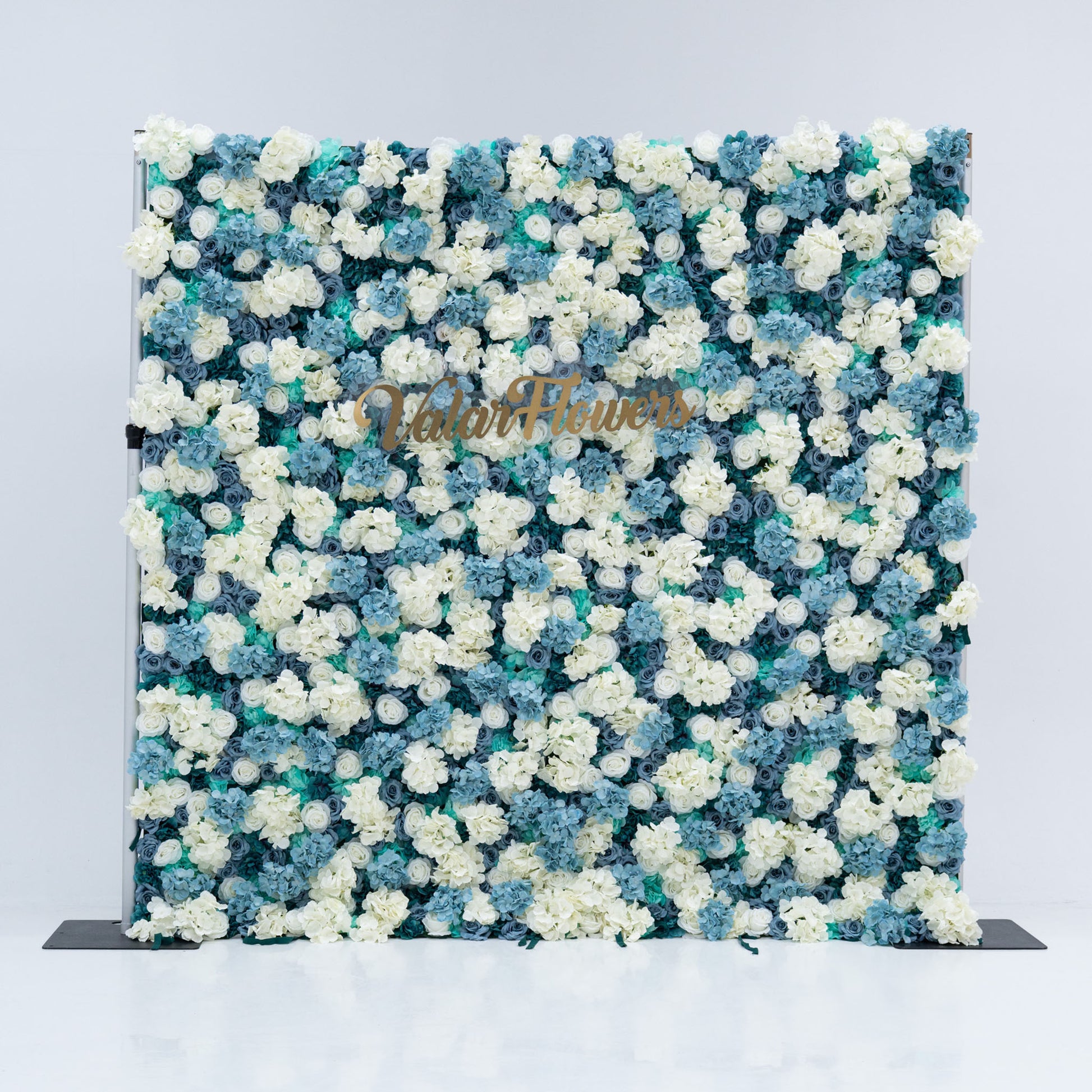 This photo showcases a stunning floral backdrop by Valar Flowers, featuring a dense arrangement of artificial roses in white and varying shades of blue. The backdrop is set against a plain white background, creating a clean and elegant look. The gold lettering of "Valar Flowers" adds a touch of sophistication, making it an ideal decorative piece for weddings, photo shoots, or other special events. The roses are meticulously arranged to create a visually appealing pattern, enhancing the overall aesthetic of 