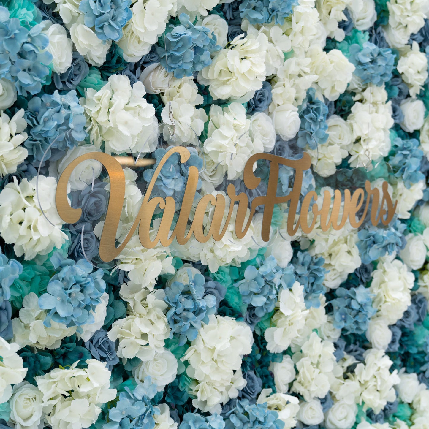 This photo showcases a stunning floral backdrop by Valar Flowers, featuring a dense arrangement of artificial roses in white and varying shades of blue. The backdrop is set against a plain white background, creating a clean and elegant look. The gold lettering of "Valar Flowers" adds a touch of sophistication, making it an ideal decorative piece for weddings, photo shoots, or other special events. The roses are meticulously arranged to create a visually appealing pattern, enhancing the overall aesthetic of 