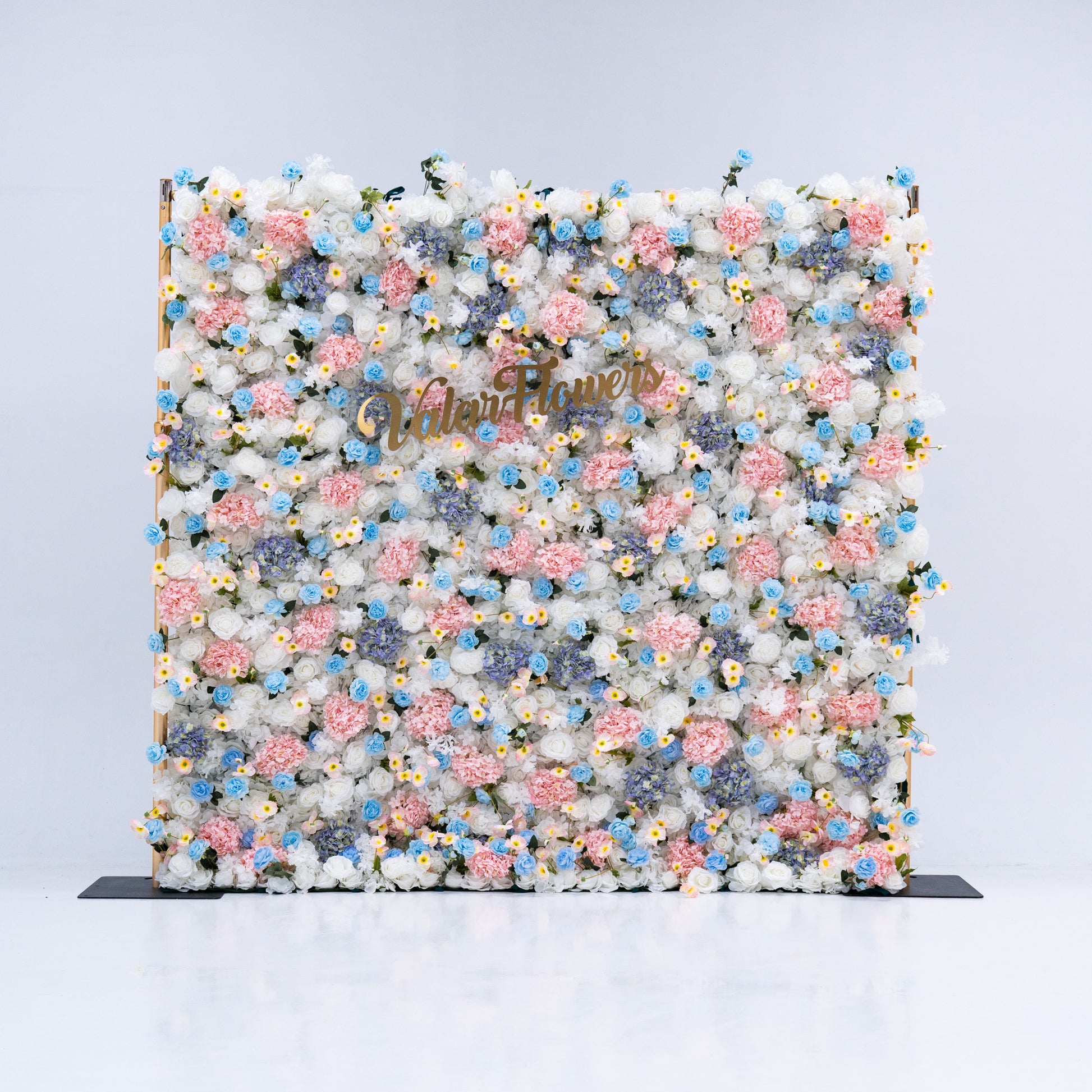 This photo features a stunning floral backdrop from Valar Flowers, showcasing a dense arrangement of artificial flowers in various shades of pink, blue, and white. The gold lettering of "ValarFlowers" adds a touch of elegance, making it an ideal decorative piece for weddings, photo shoots, or other special events. The meticulous arrangement of the flowers enhances the overall aesthetic, adding a touch of natural beauty and charm to any setting.