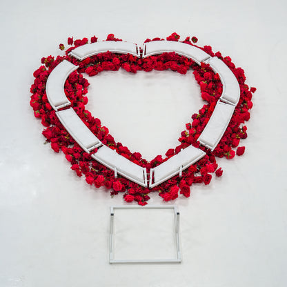 A heart - shaped structure with red roses adorning its edges, partially assembled with white frame pieces visible. Below it lies a square white metal frame. The vibrant red blooms contrast with the white components, suggesting a decorative floral installation in progress. 